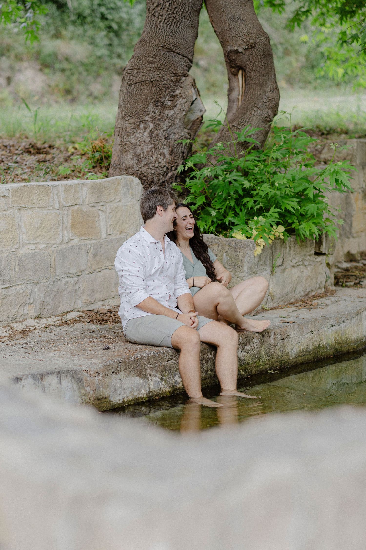 Couple sitting on stone steps by a serene pond, feet in water, surrounded by lush greenery and a large tree.