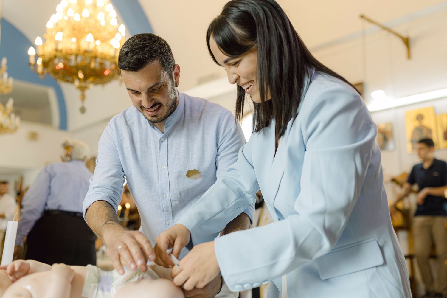Man and woman in formal attire performing a baby baptism ceremony in an elegant church setting.