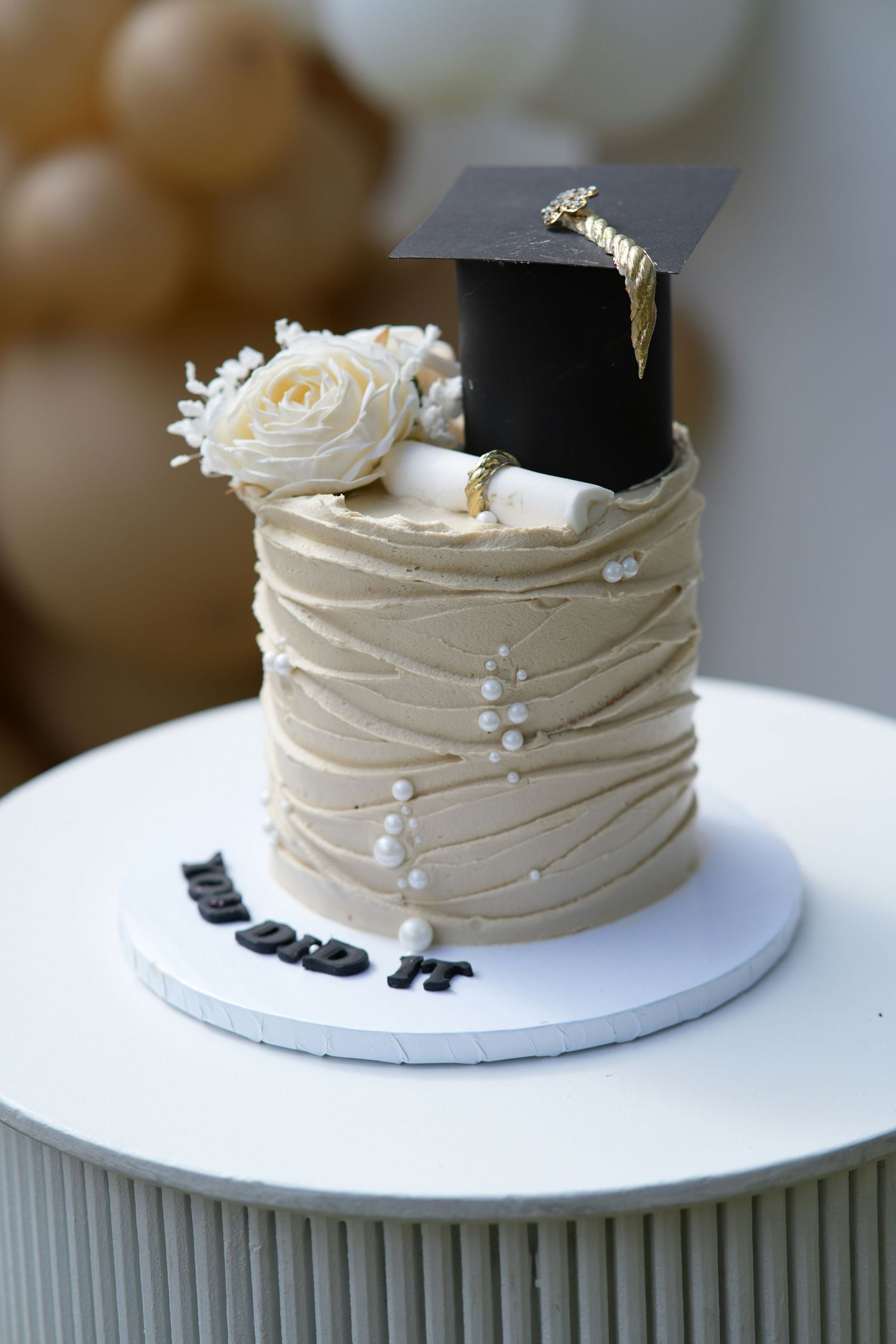 Elegant graduation cake with beige frosting, pearl details, white roses, diploma, and mortarboard topper reading You Did It.