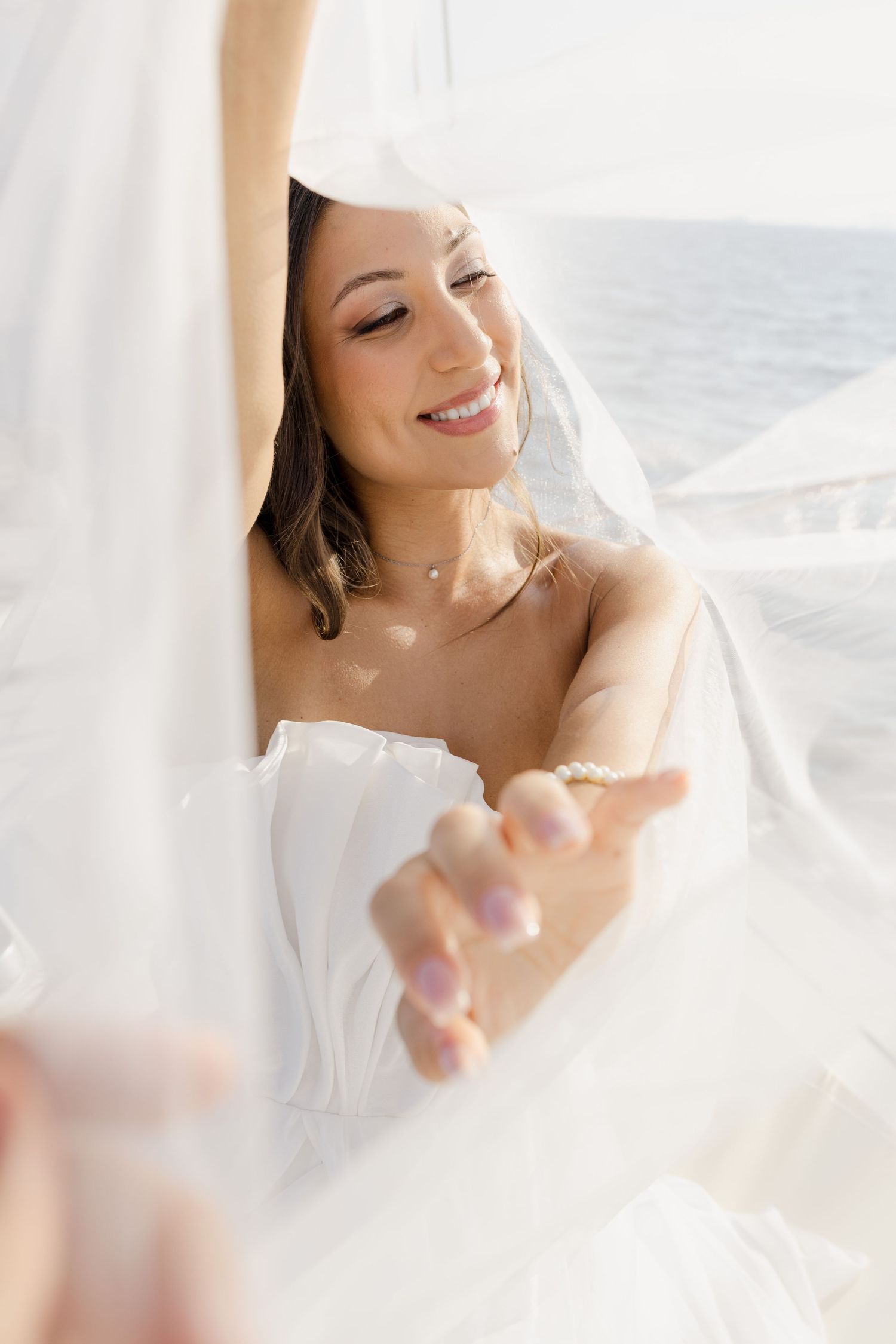 Smiling bride in white strapless gown and veil reaching out her hand near the ocean on a sunny day.