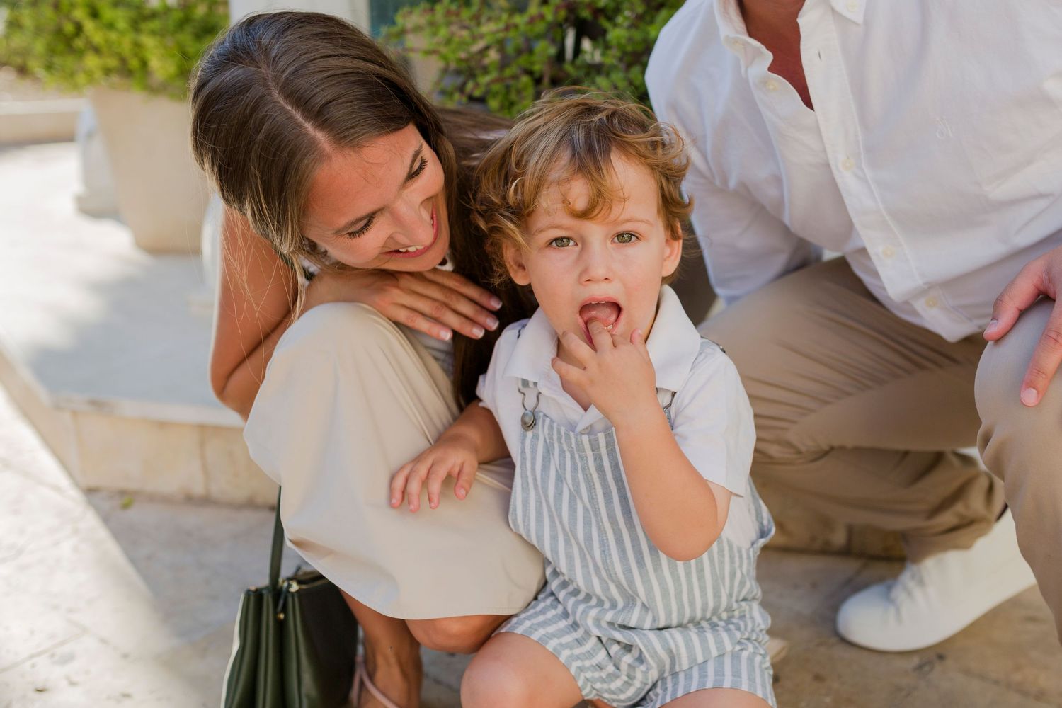A woman hugs a curly-haired toddler boy outdoors while a man crouches beside them in bright sunlight.