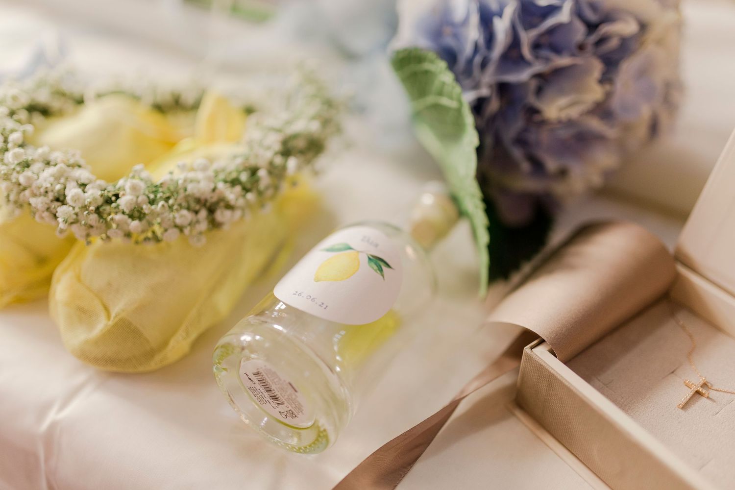 Wedding flat lay with floral crown, lemon perfume bottle, hydrangea bouquet, and gift box on white surface.