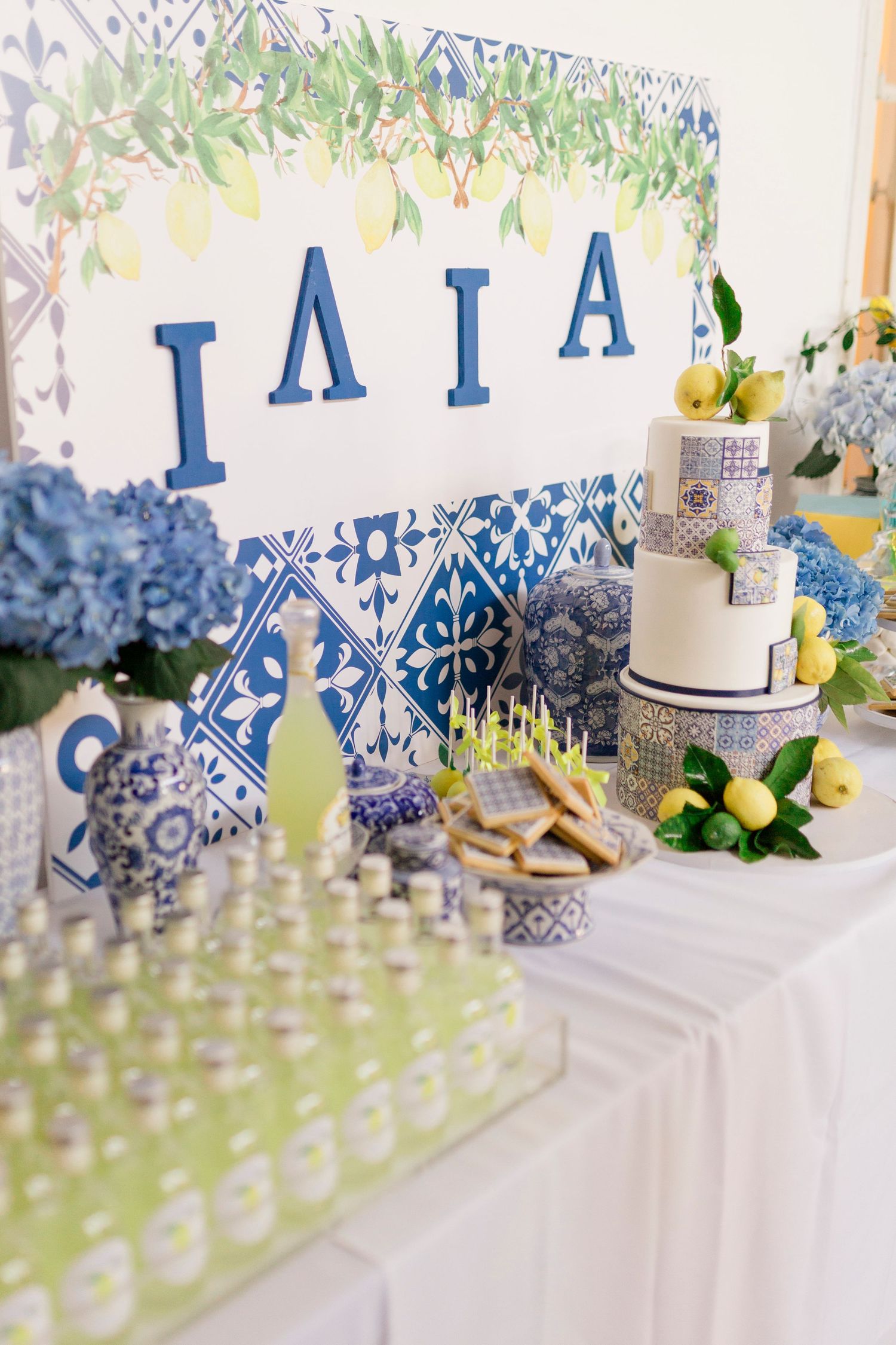 Blue and white Mediterranean-themed birthday dessert table with tiered cake, lemons, hydrangeas, and decorative tiles.