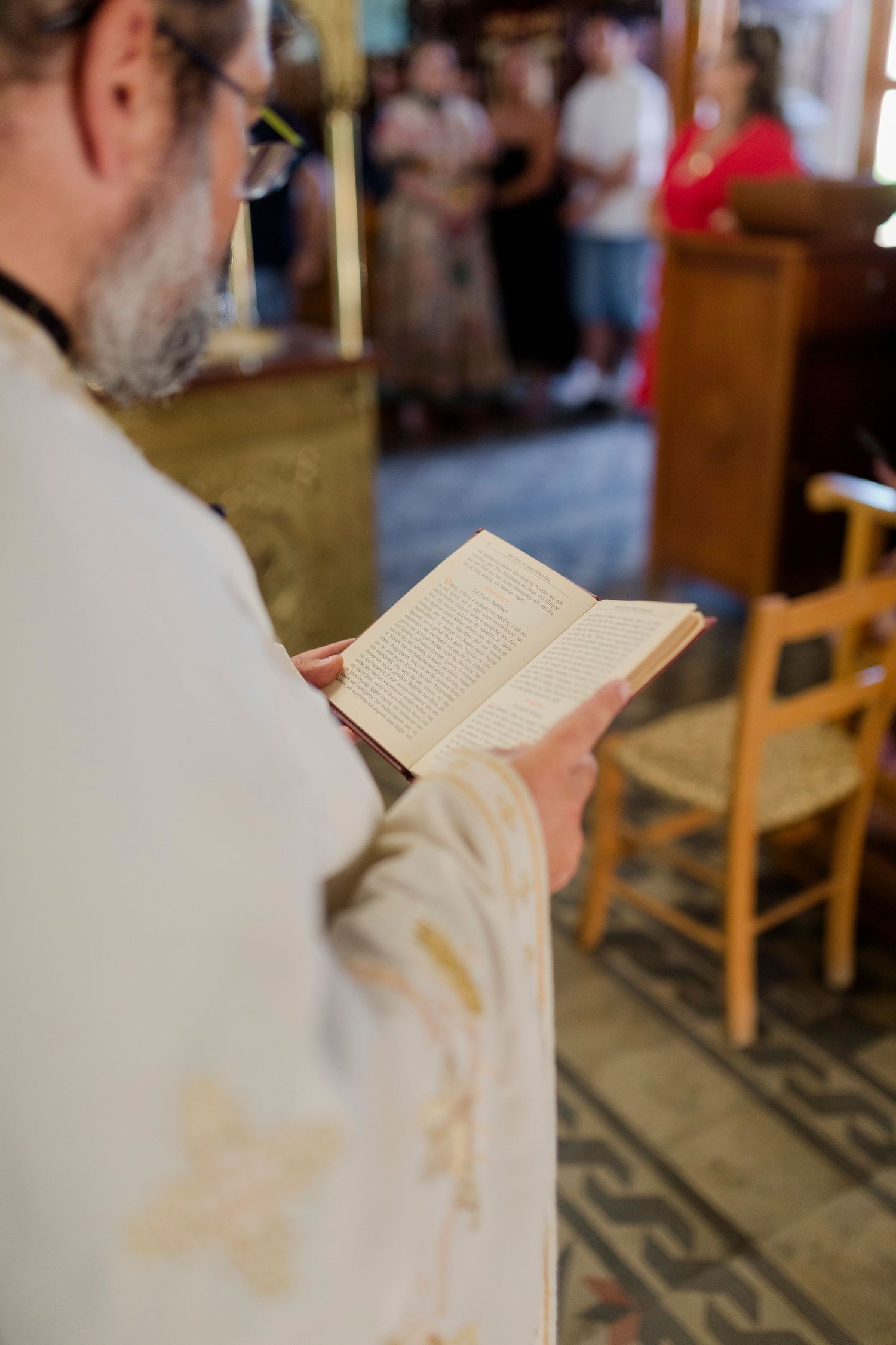 Orthodox priest in white vestments reading a prayer book during a church ceremony with congregation in background.