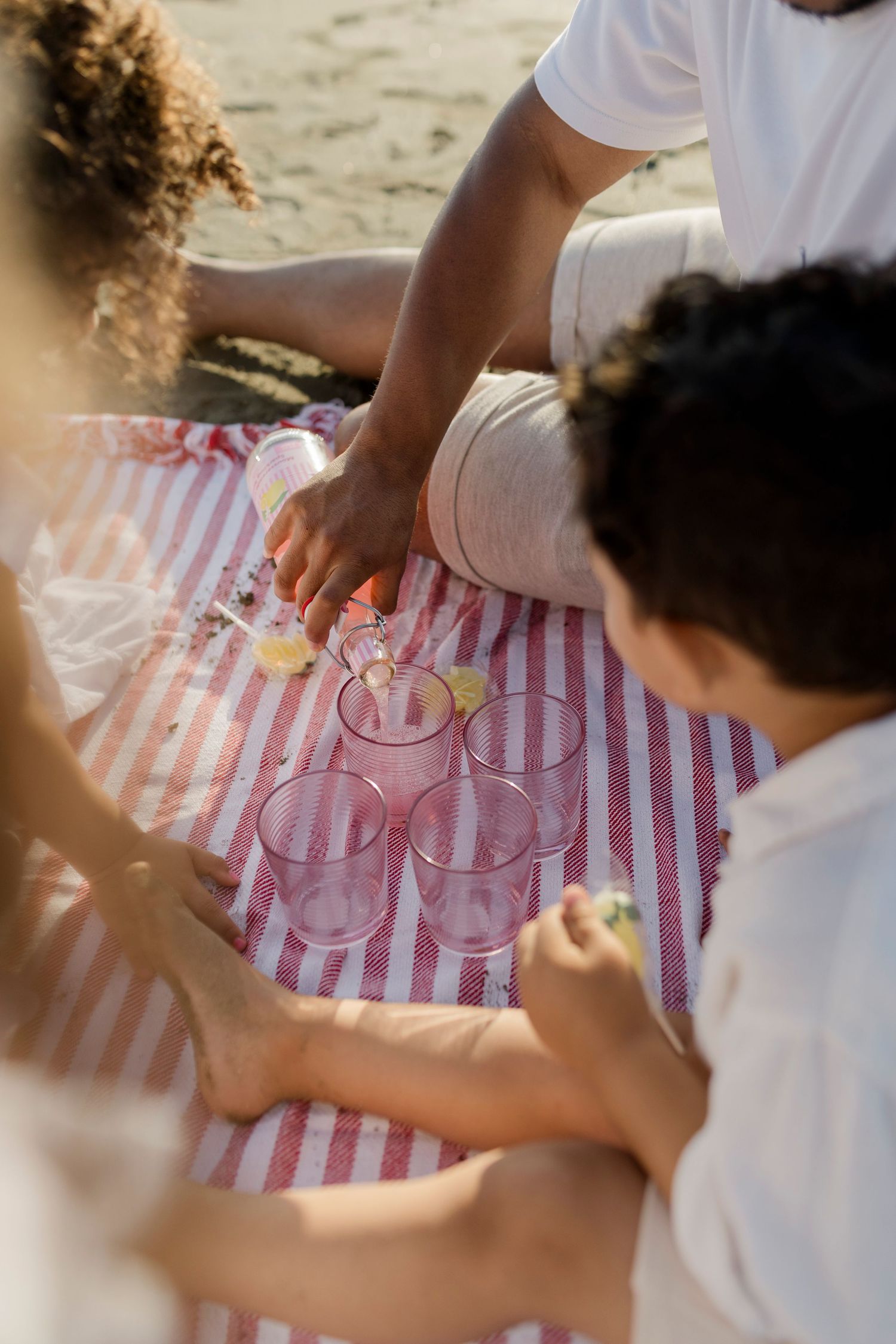 Friends enjoying a beach picnic on a striped blanket with pink plastic cups and snacks on a sunny day.