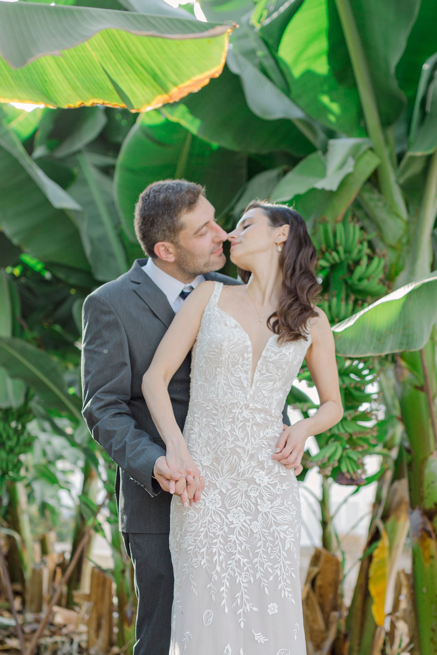 Bride and groom sharing a romantic kiss surrounded by lush tropical banana trees in an outdoor wedding portrait.