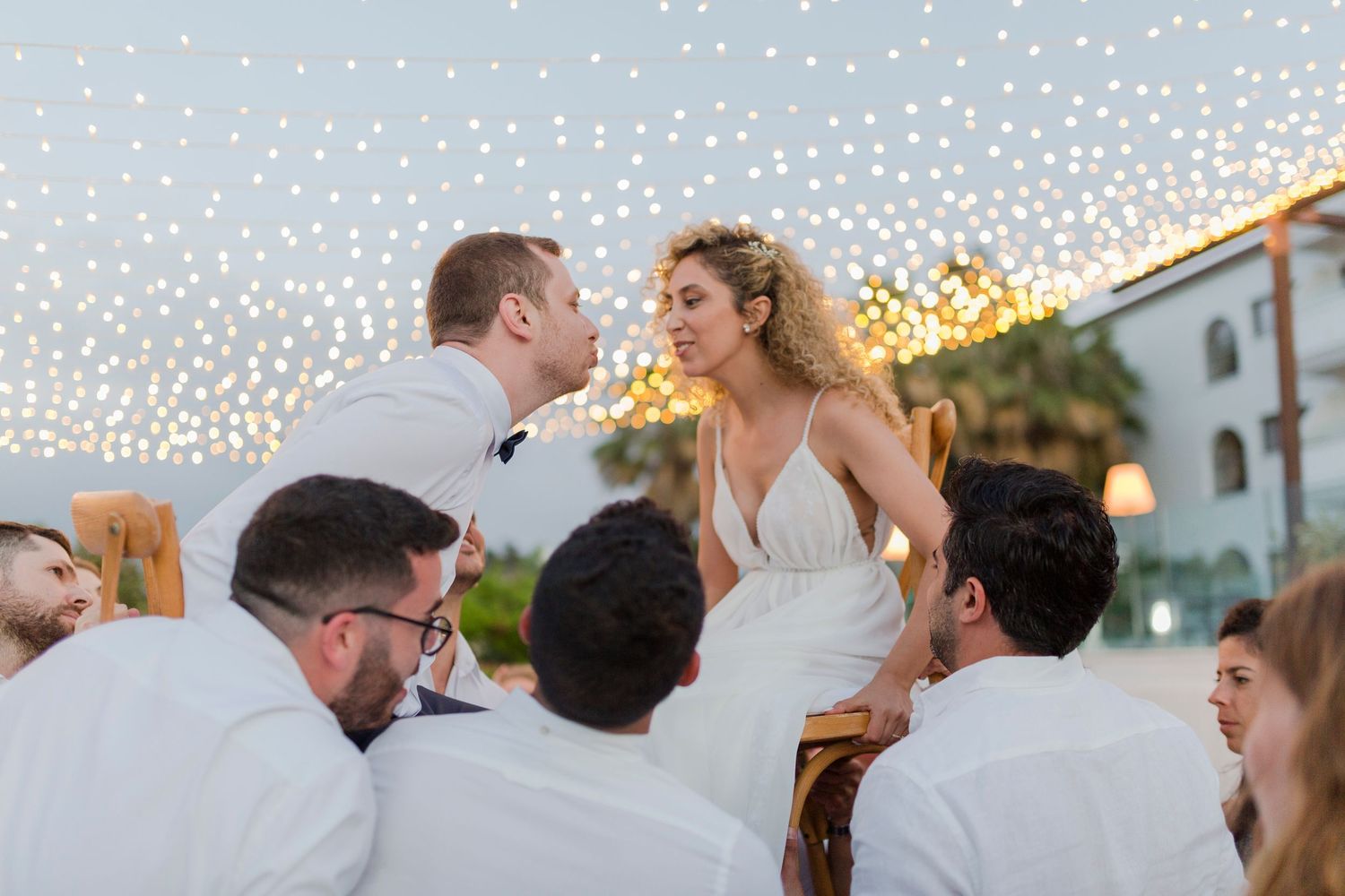 Bride and groom share a romantic moment surrounded by guests under twinkling fairy lights at reception.
