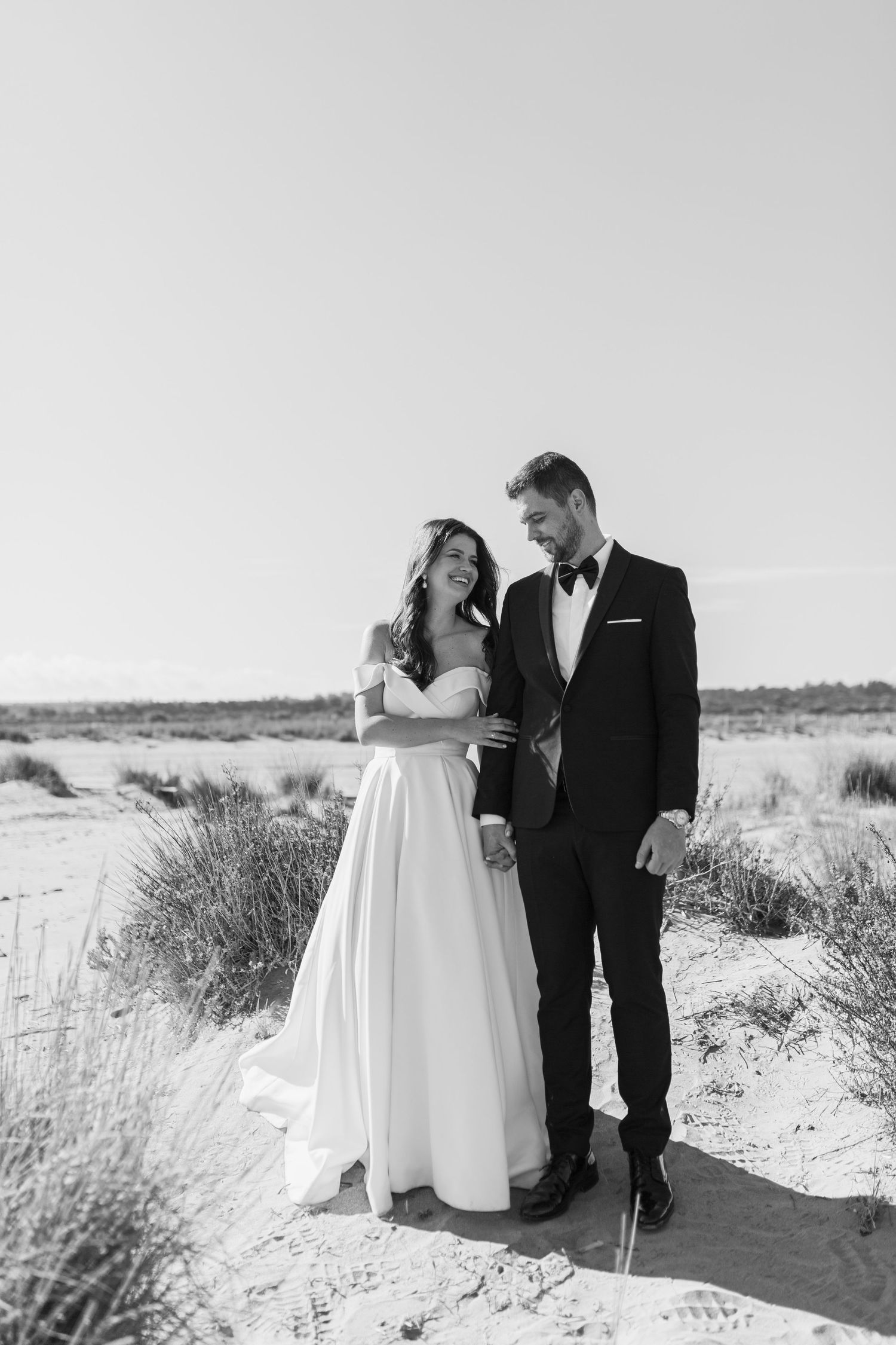 Black and white photo of bride and groom holding hands on sandy landscape, bride in flowing off-shoulder gown.