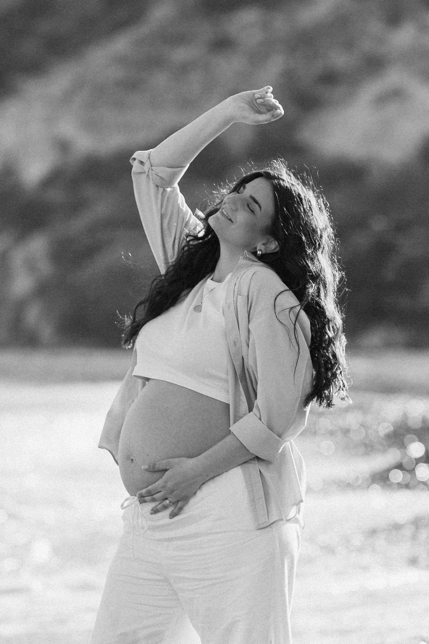 Black and white photo of a pregnant woman in white outfit posing outdoors, cradling her belly with one arm raised gracefully.