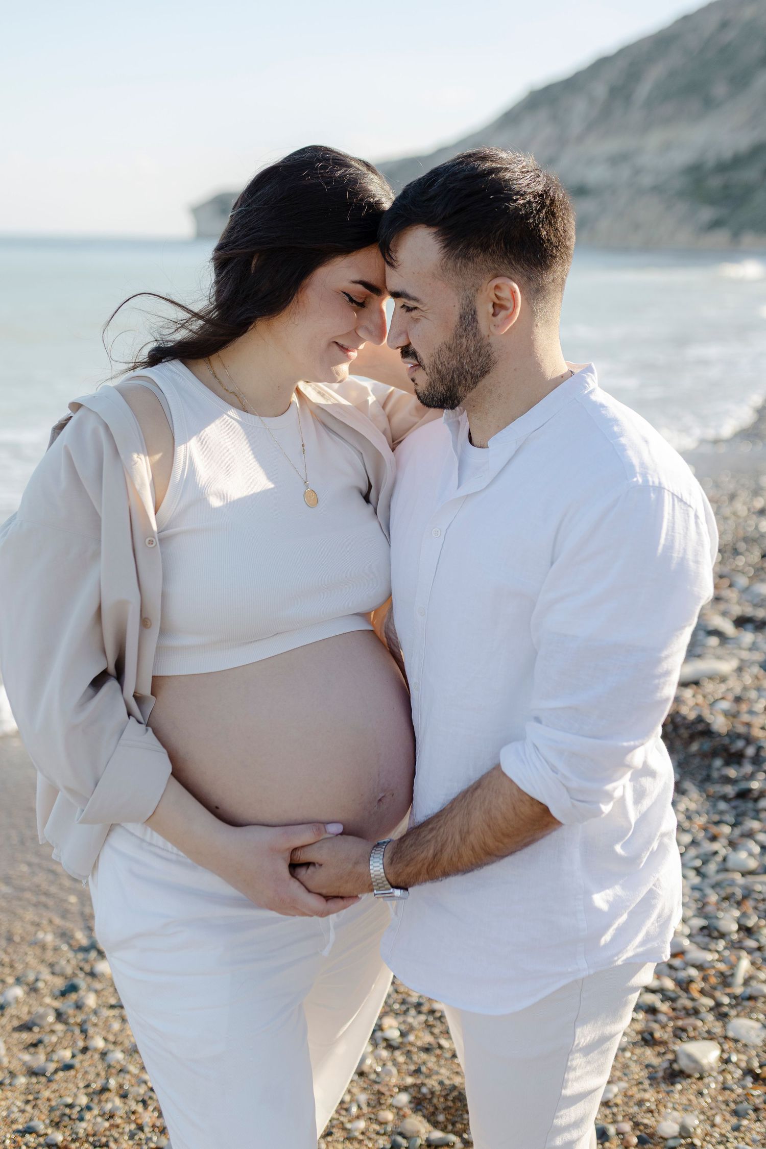Pregnant couple in white clothing tenderly touching foreheads on a rocky beach by the sea.