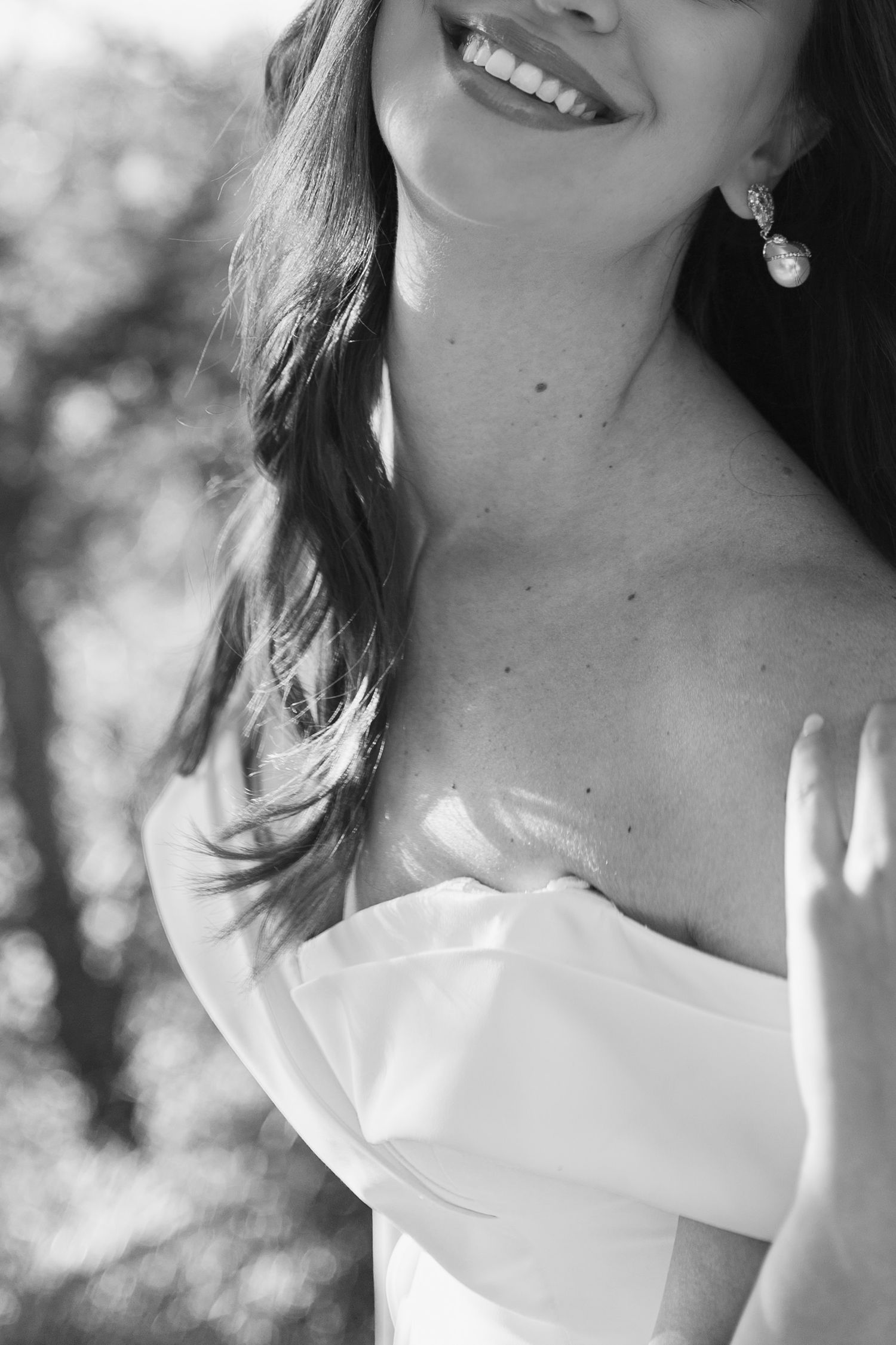 Black and white portrait of smiling bride in off-shoulder gown wearing pearl drop earrings outdoors.