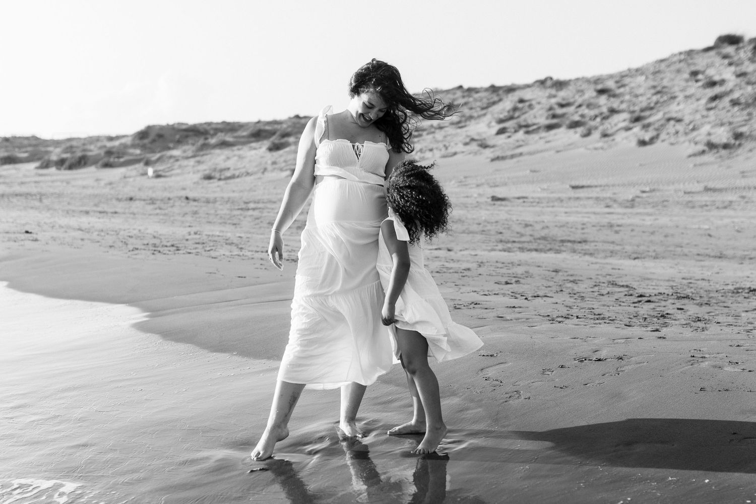 Black and white beach photo of pregnant woman in white dress walking with young child by ocean waves.