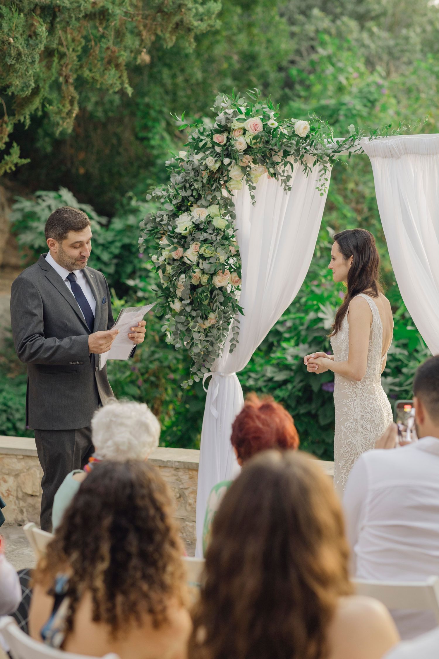 Outdoor wedding ceremony with groom reading vows to bride under floral arch with white draping and greenery.