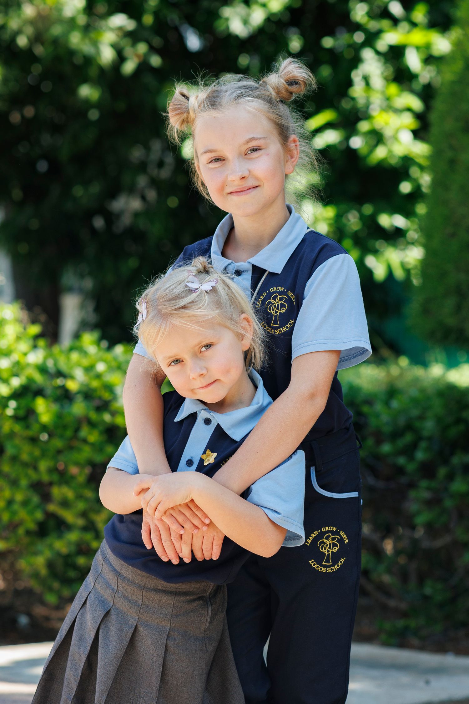 Two young girls in matching school uniforms embrace outdoors, smiling amid lush green foliage on a sunny day.