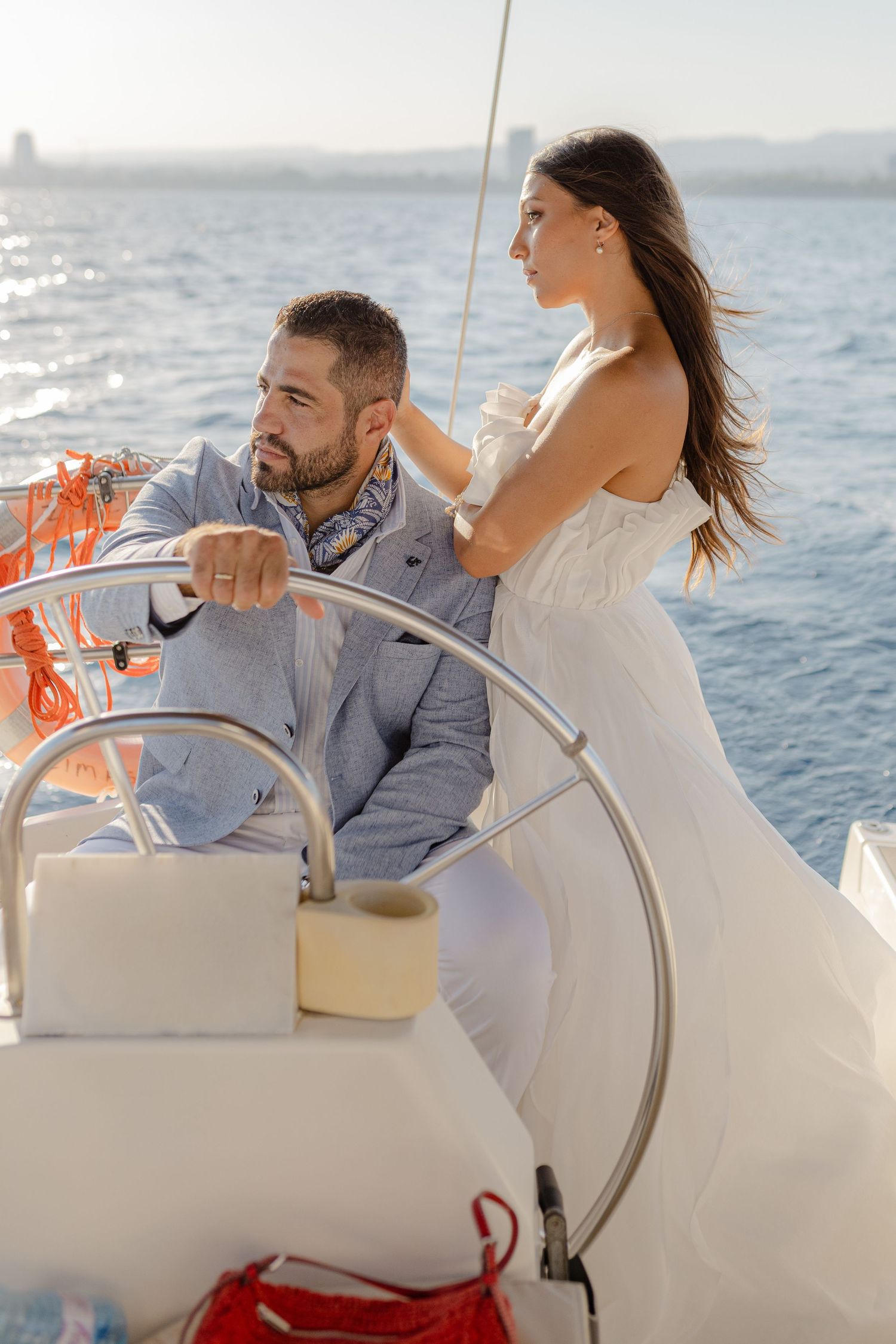 Elegant couple on a sailboat, man in suit at helm, woman in white dress, blue ocean background.