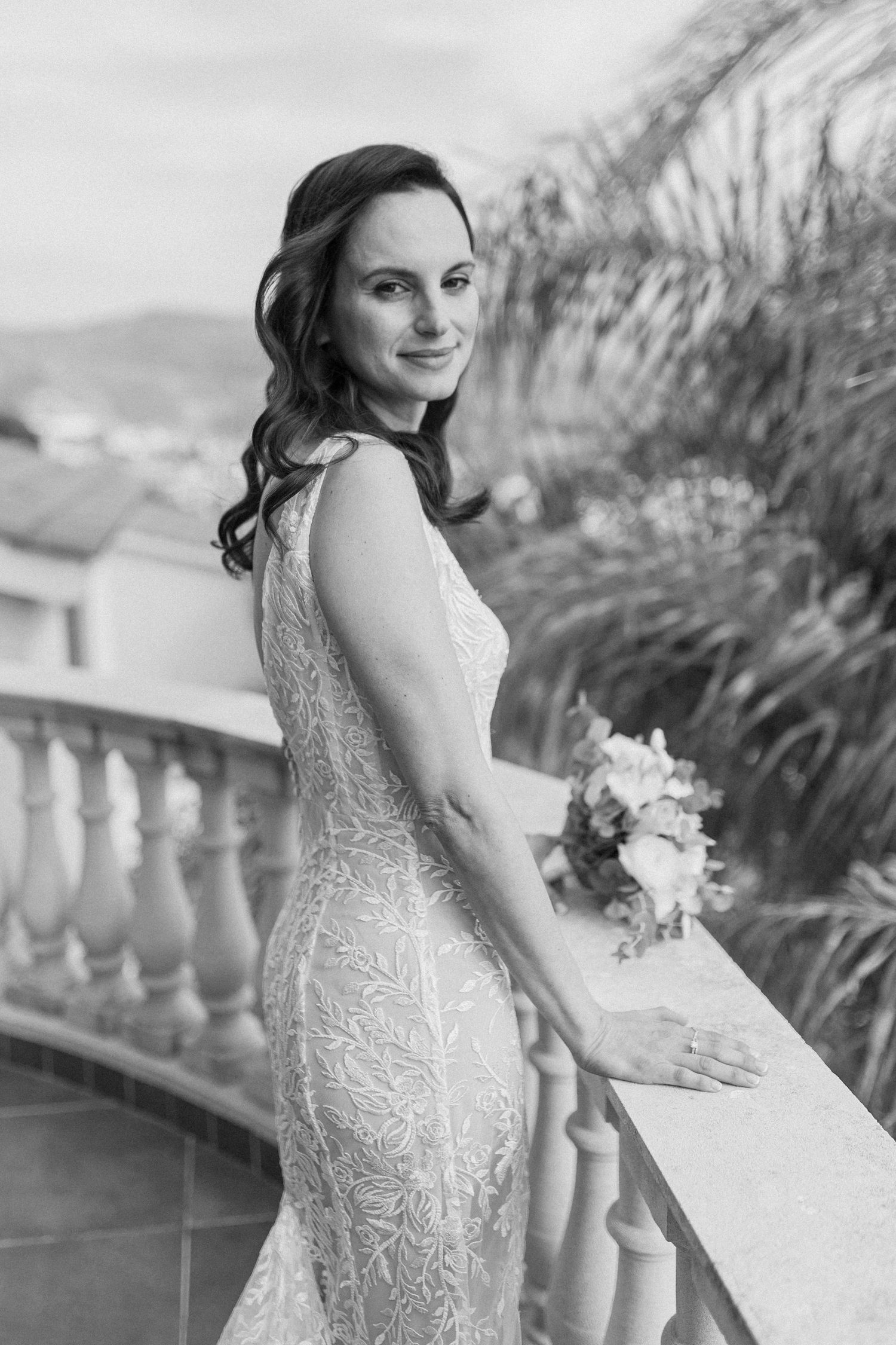 Elegant bride in lace wedding gown smiling over shoulder while holding bouquet on balcony in black and white.