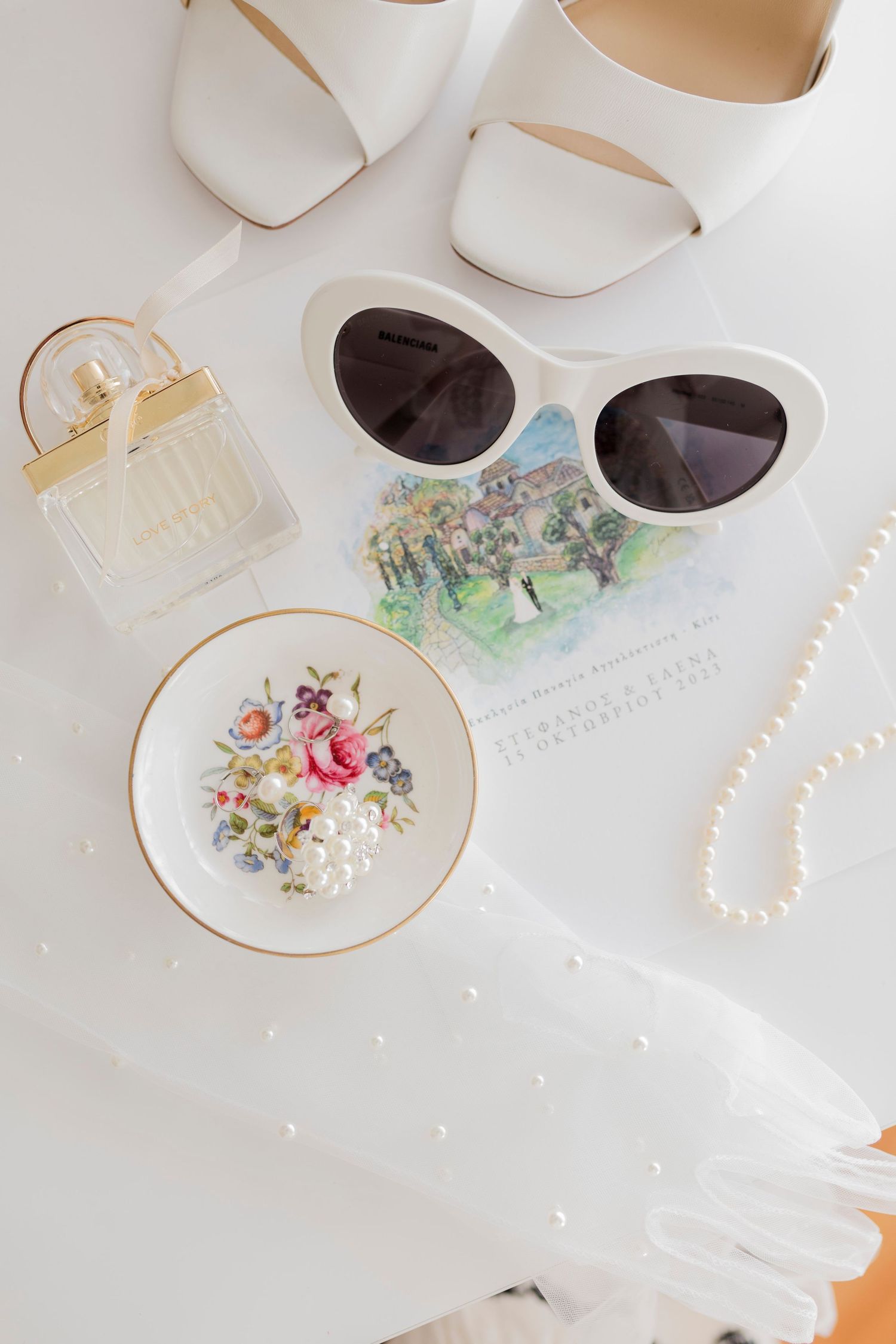 Elegant bridal flat lay featuring white heels, cat-eye sunglasses, pearl necklace, perfume, floral dish, and wedding invitation.