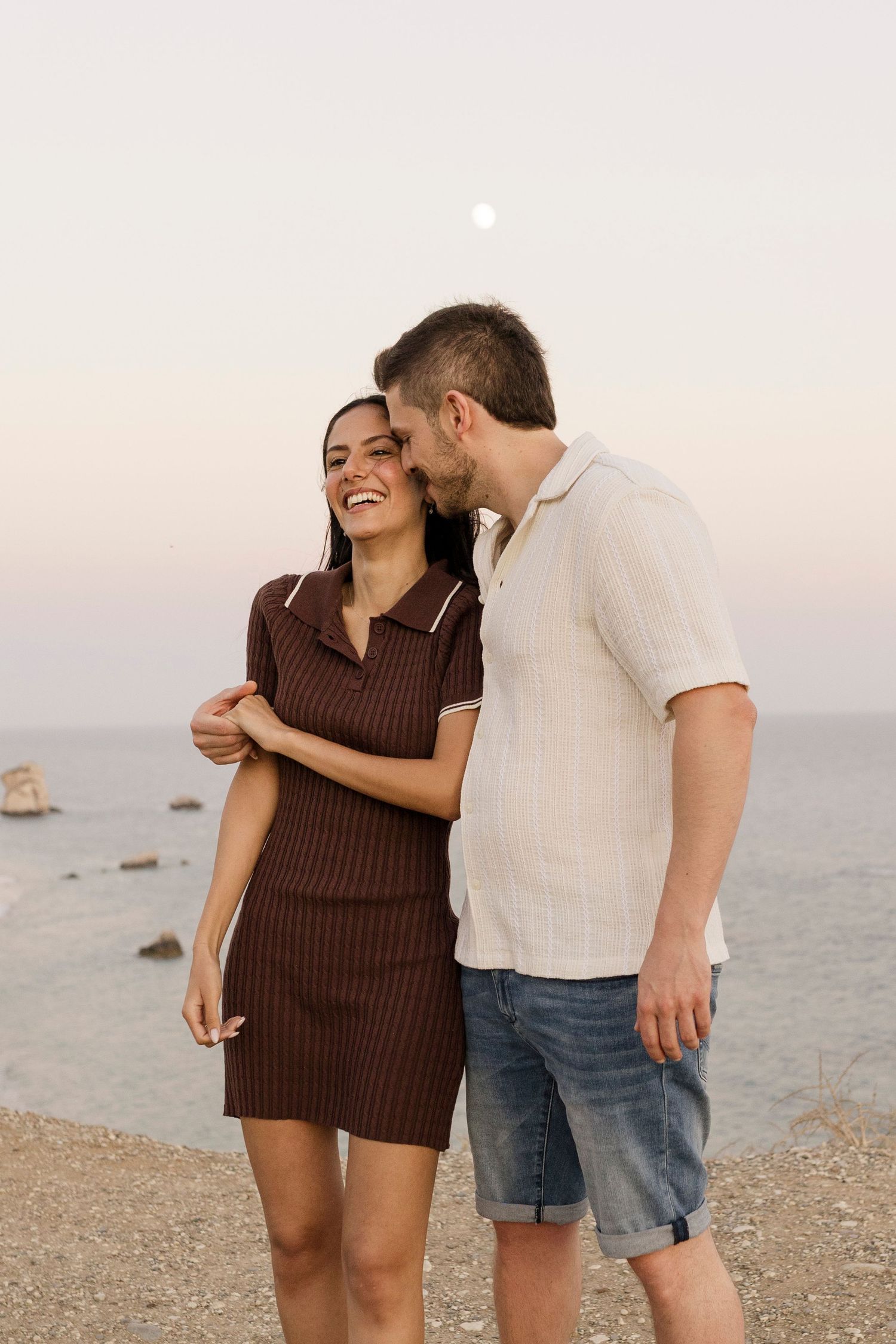 Couple embracing and laughing on a rocky beach at sunset with the moon visible in the pastel sky.