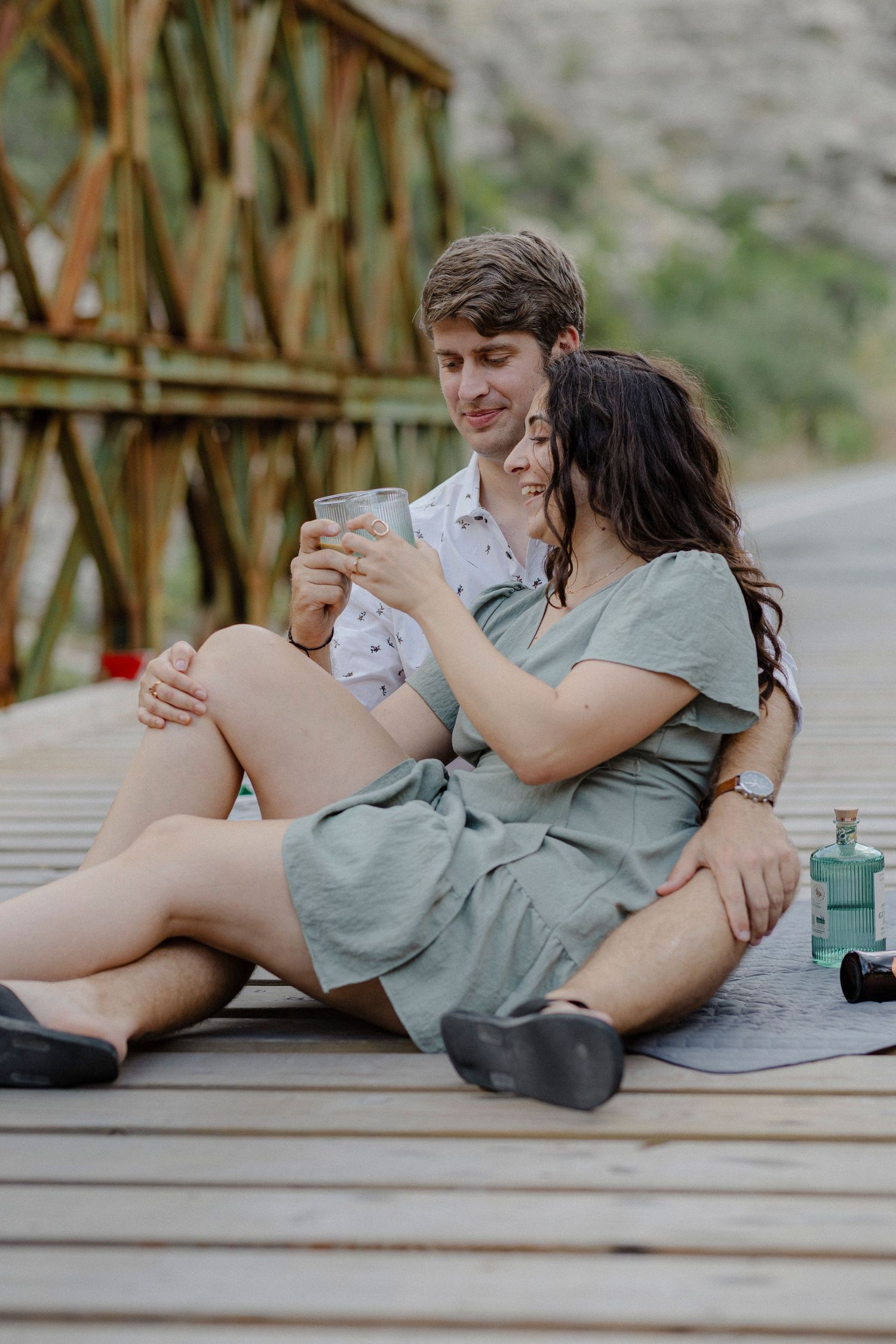 Couple sitting on wooden bridge deck sharing a drink, woman in sage dress leaning against man outdoors.