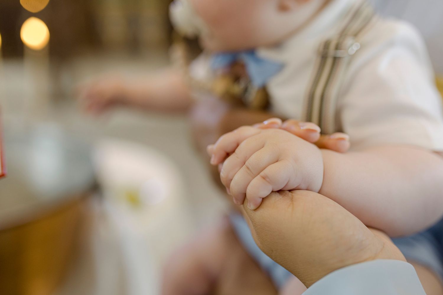 Close-up of a baby grasping an adult's hand, wearing a white outfit with a blue bow at a christening.