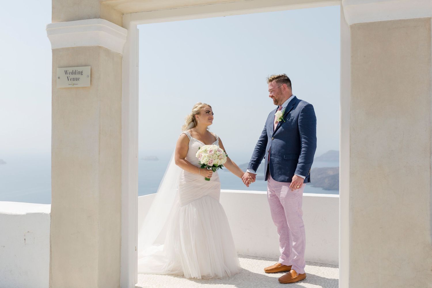 Bride and groom hold hands under white archway with Santorini sea view on their wedding day.