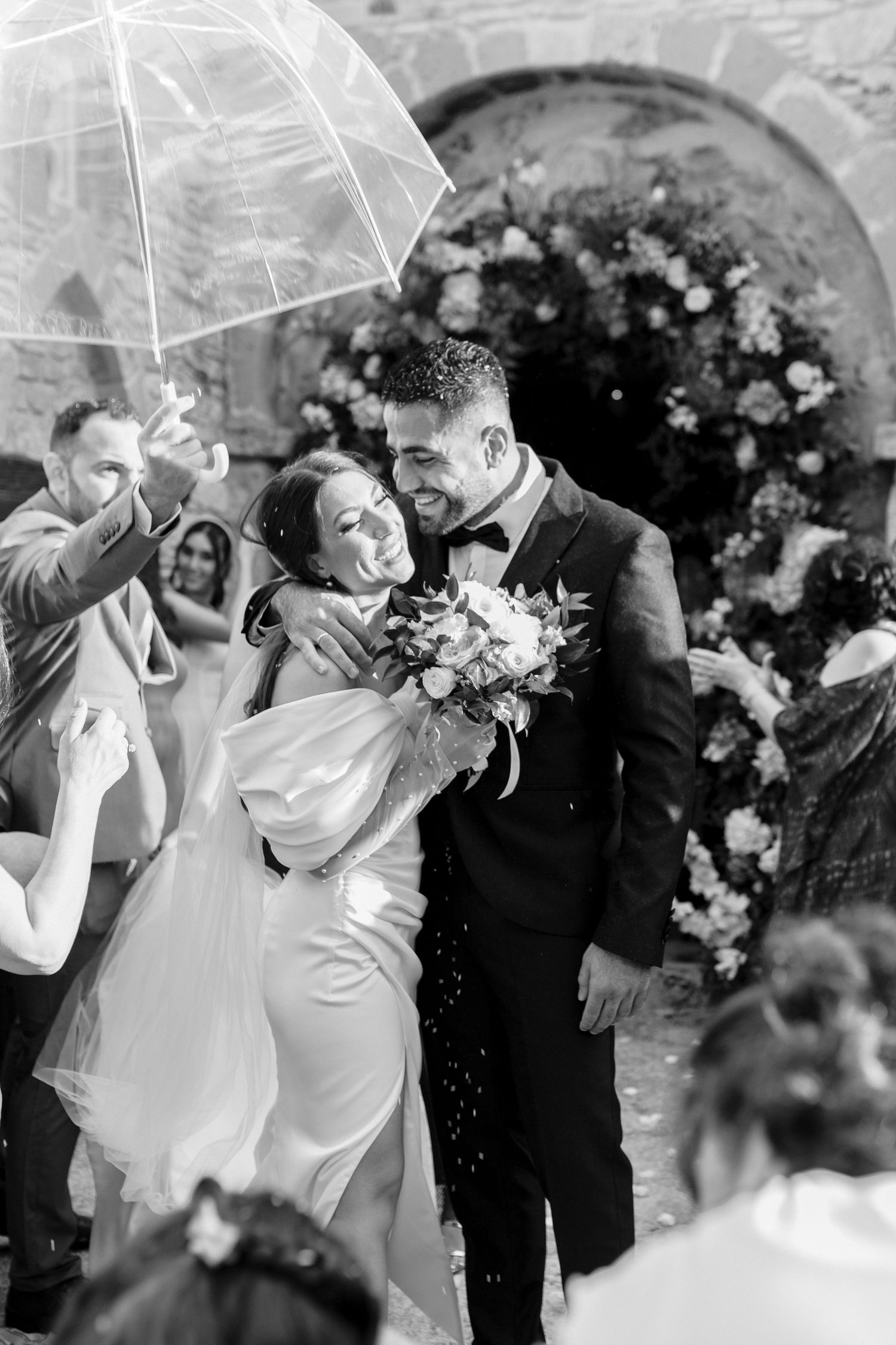 Black and white photo of newlyweds exiting ceremony under clear umbrella as guests shower them with confetti.