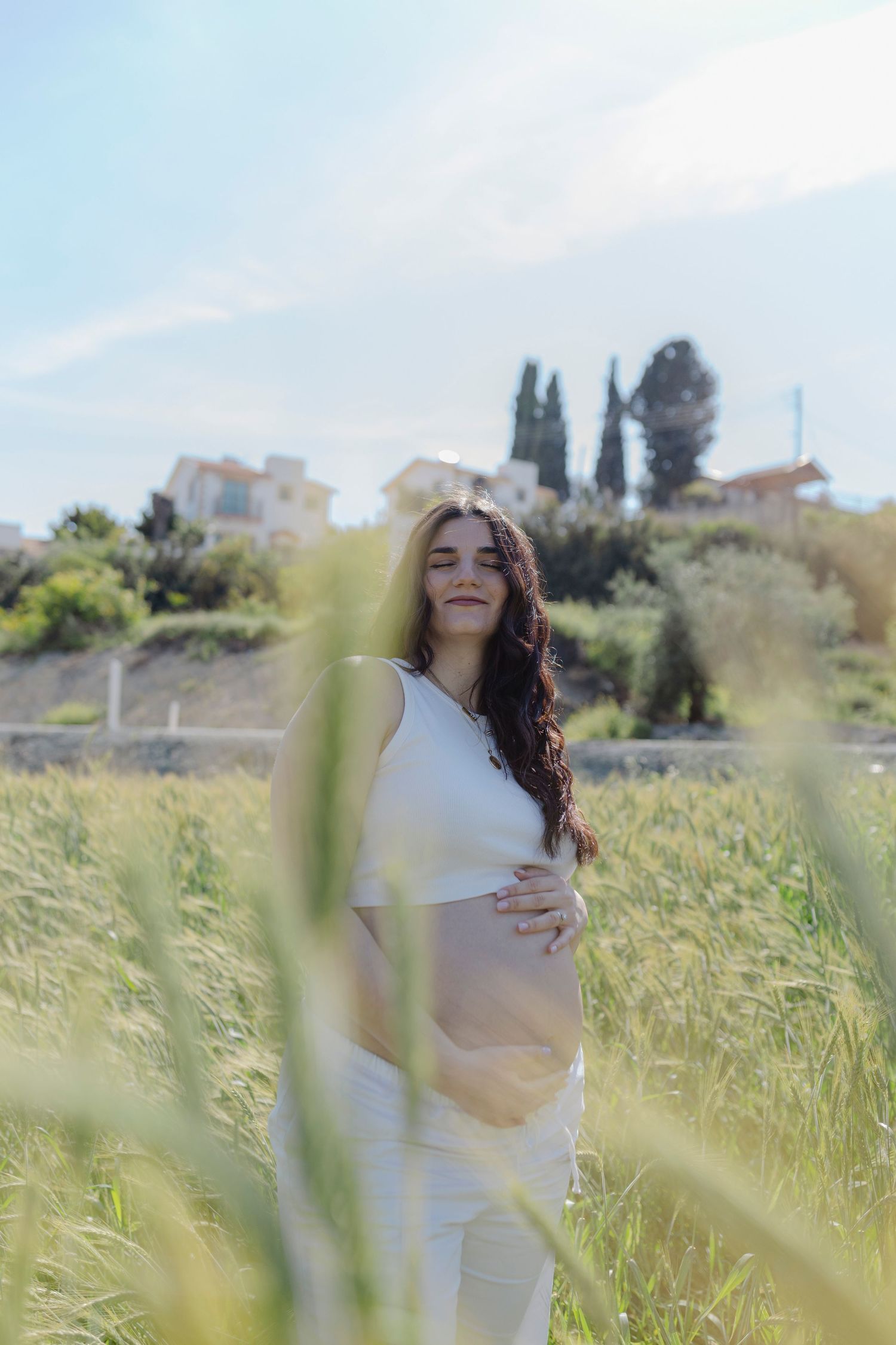 Pregnant woman in white outfit standing in tall grass field, gently holding her baby bump on a sunny day.