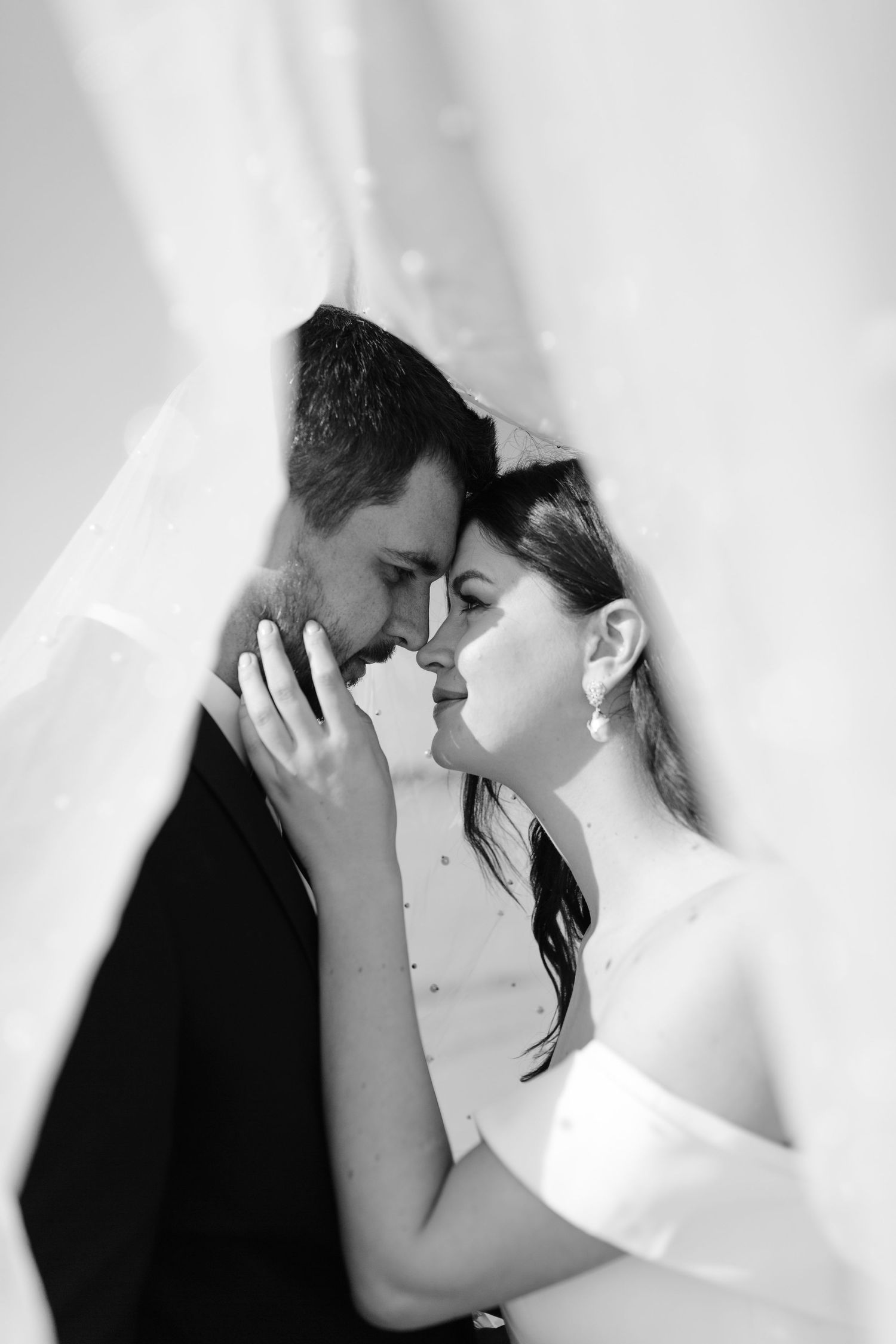 Black and white photo of bride and groom sharing an intimate moment under a wedding veil.
