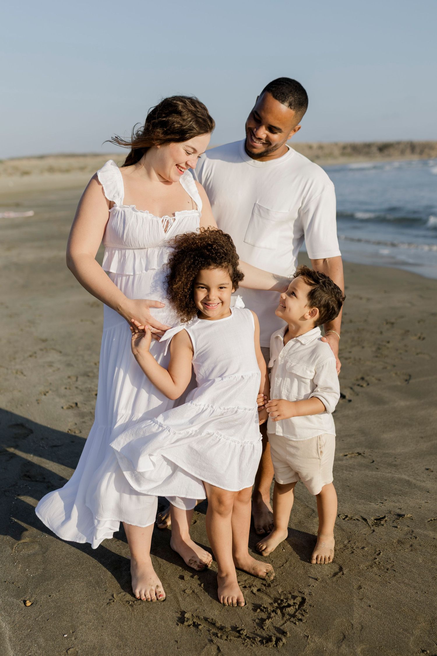 A happy interracial family of four poses on a beach in white outfits, with a pregnant mother and smiling children.