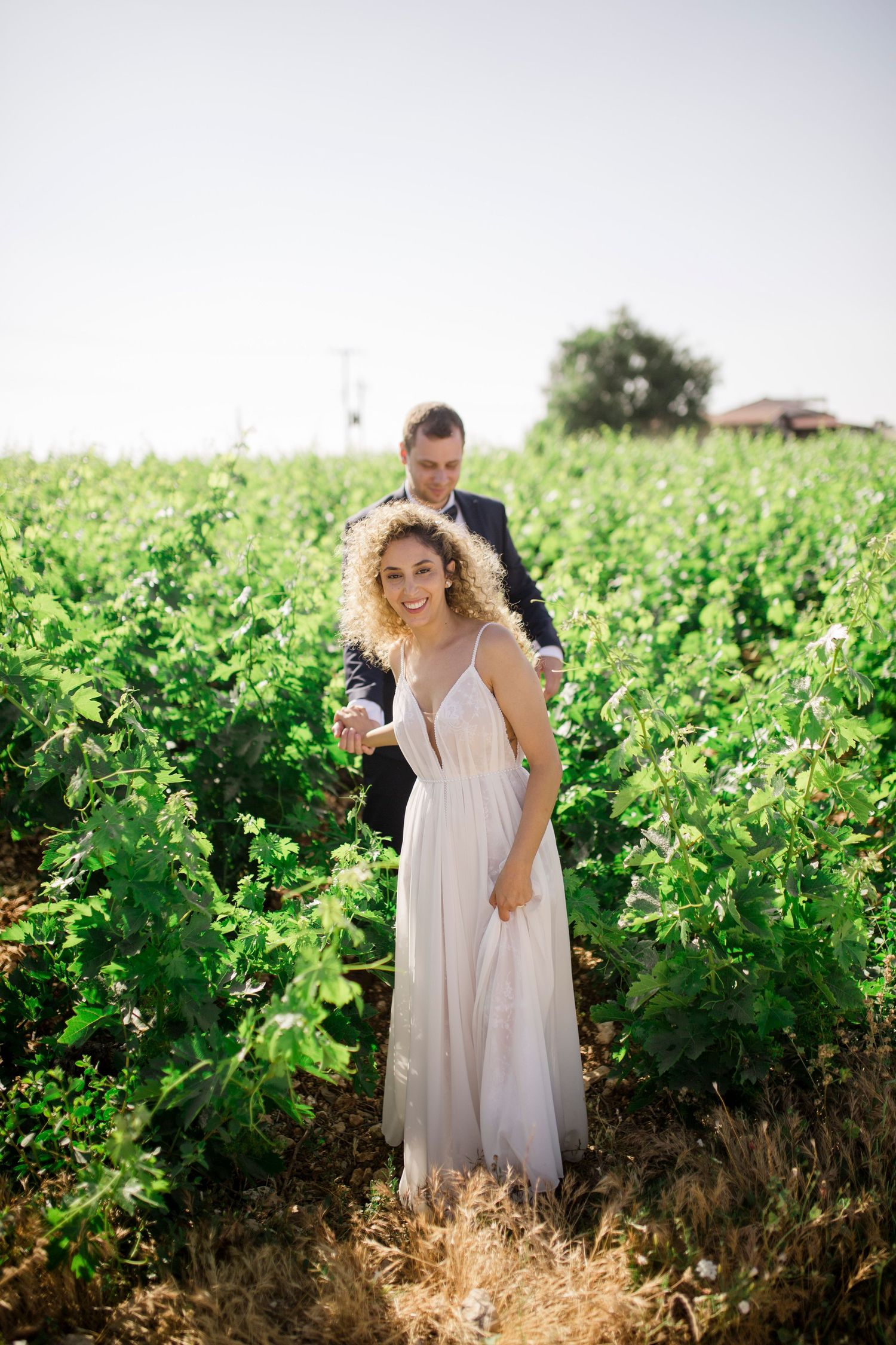 A smiling bride in a white gown and groom in a suit stand together in a lush green vineyard on a sunny day.