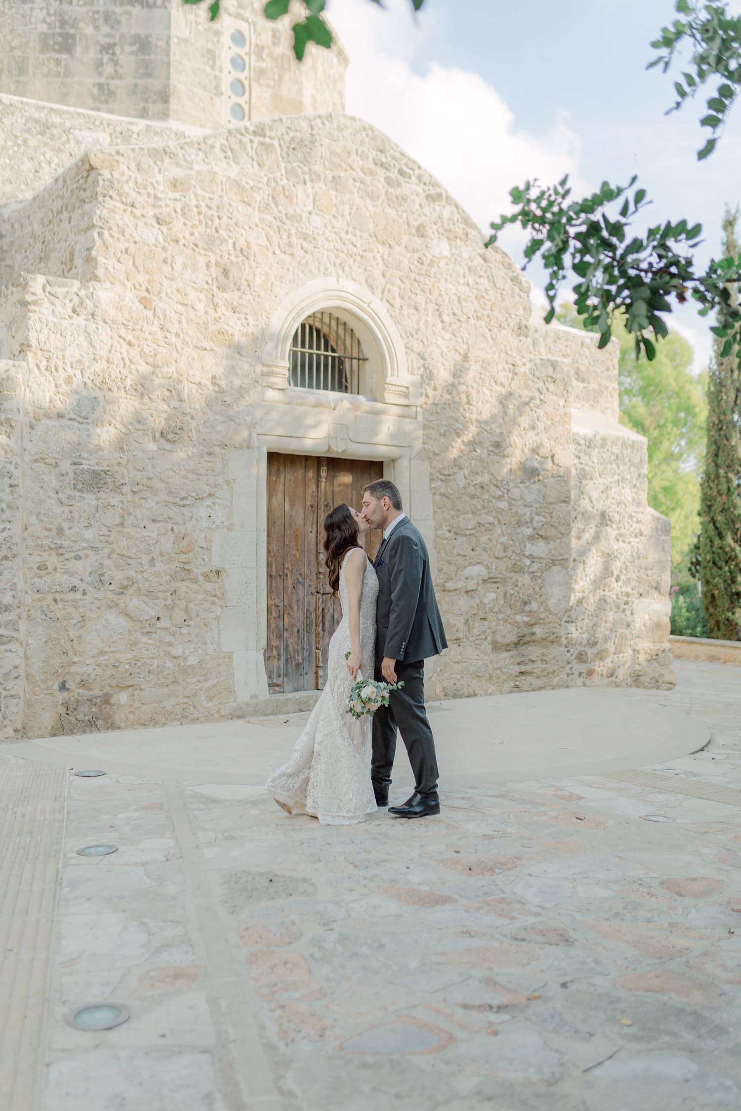 Bride and groom share a kiss outside a rustic stone church on their wedding day.