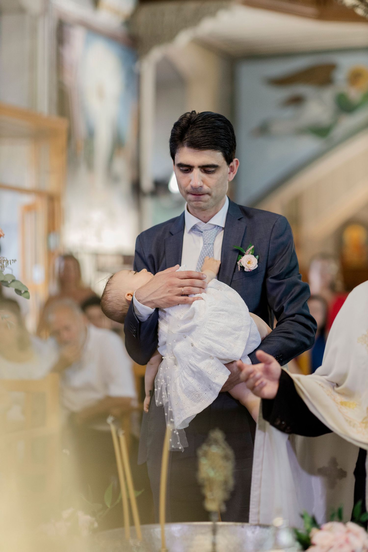 Man in suit holding infant in white christening gown during a baptism ceremony in a church.