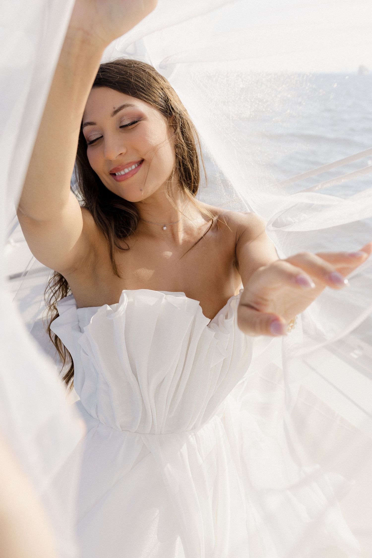 Smiling bride in strapless white dress dancing with veil flowing near ocean.