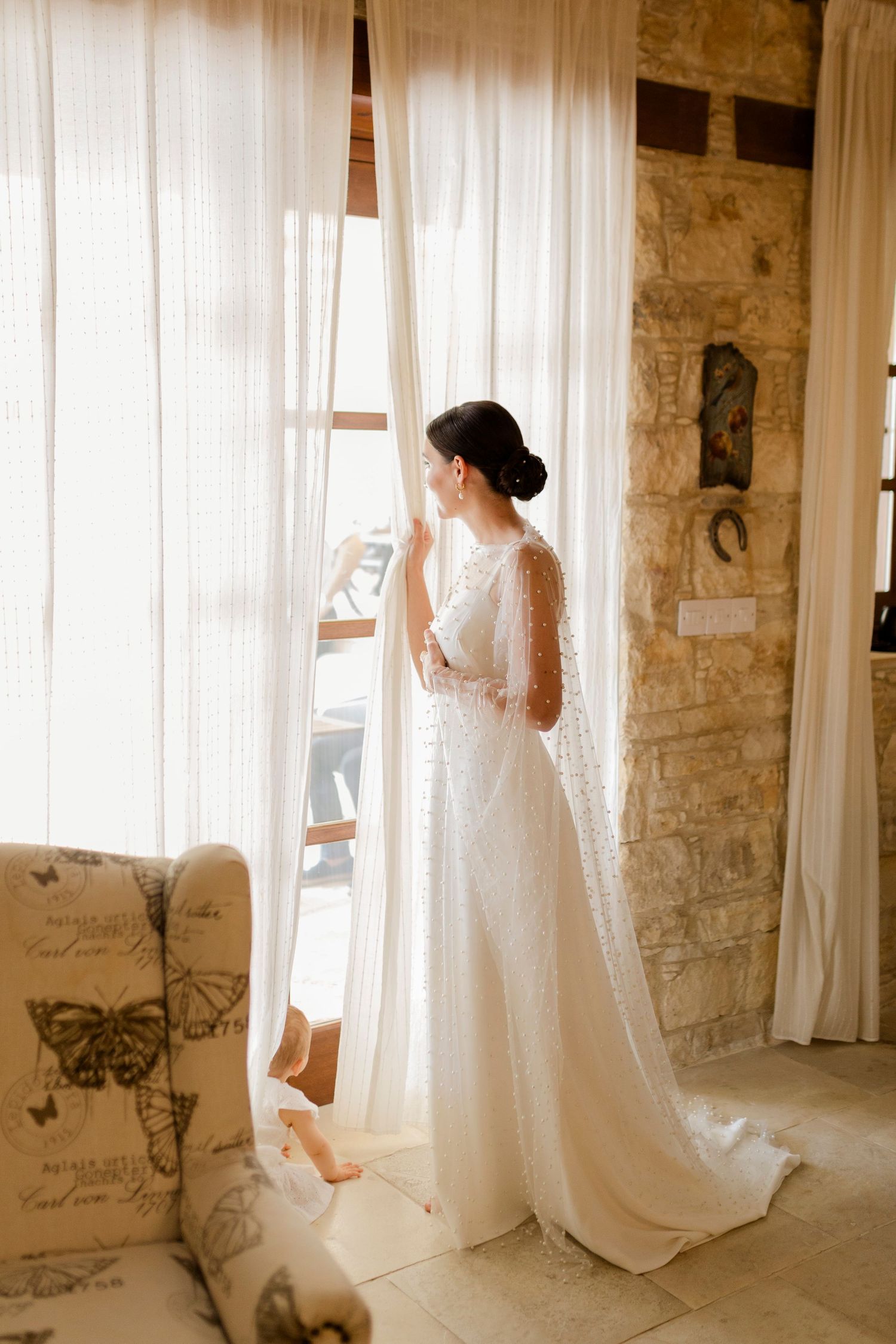 Bride in white lace wedding gown standing by sheer curtained window in stone-walled room with natural light.