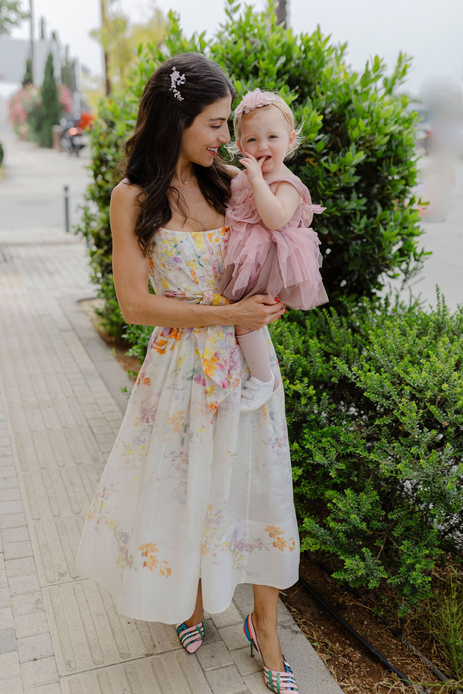 Woman in floral midi dress holds baby girl in pink tutu on a garden pathway.