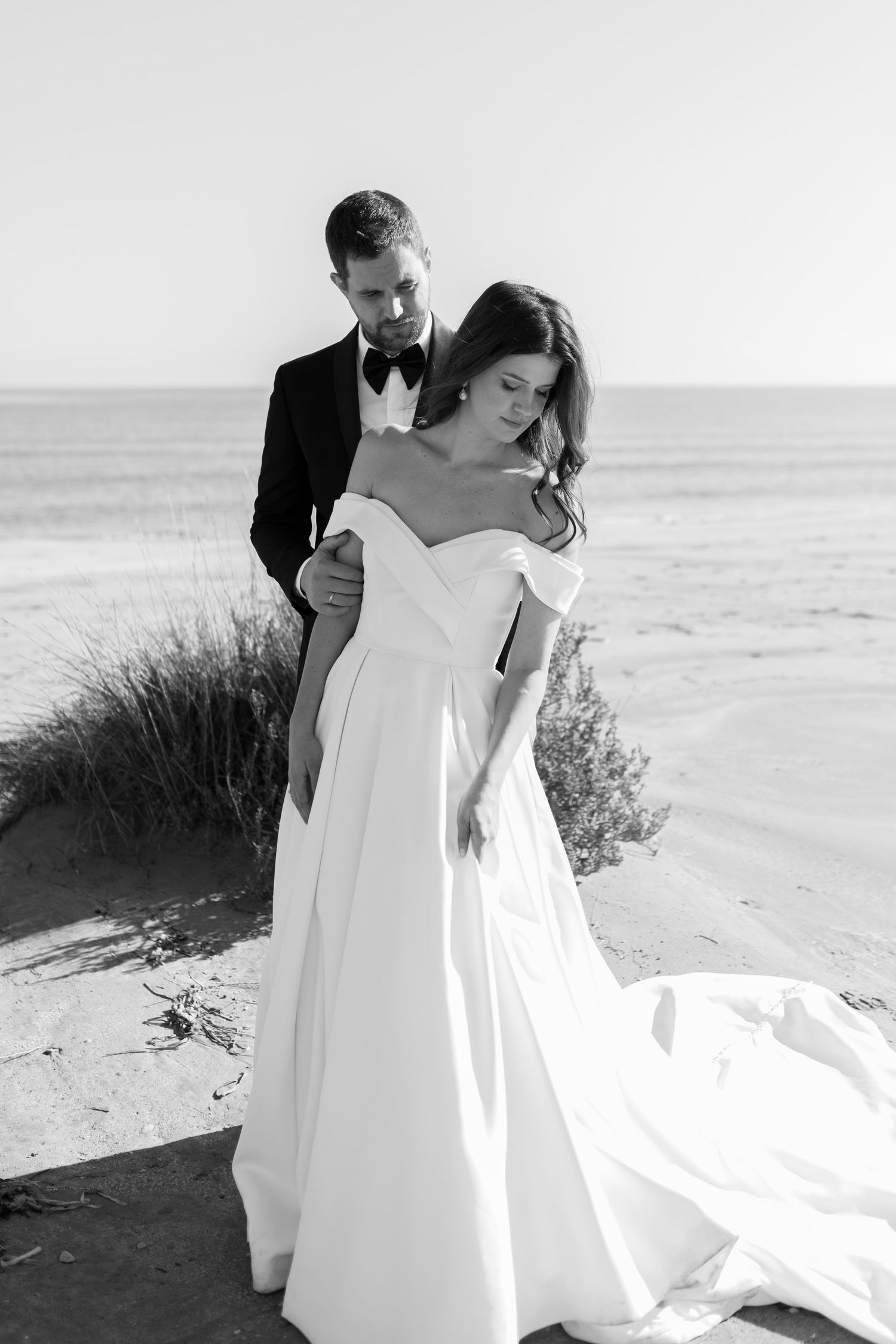 Black and white beach wedding photo of bride in off-shoulder gown embraced by groom in tuxedo.