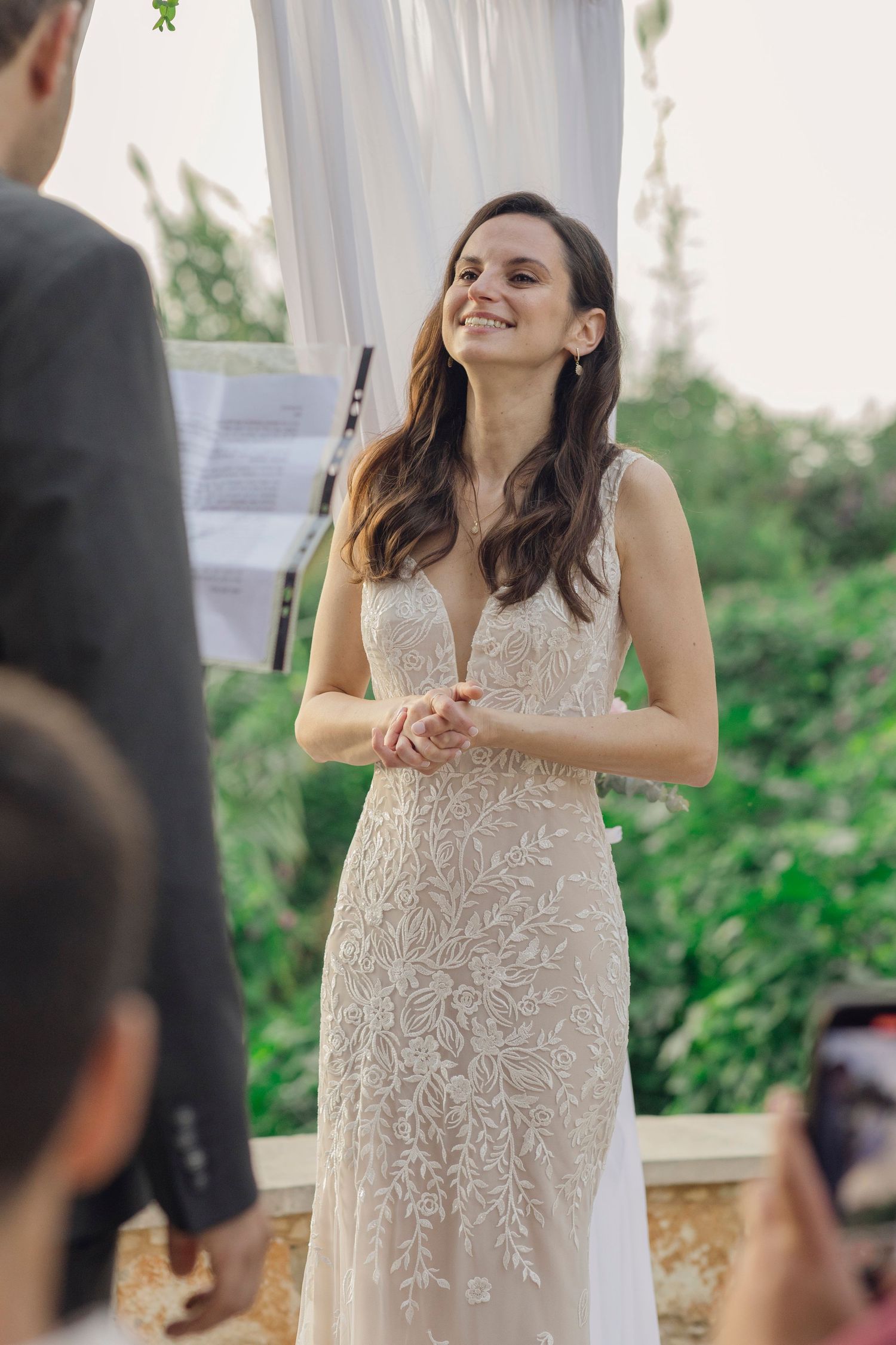 Smiling bride in embroidered lace wedding gown stands at outdoor ceremony altar with greenery backdrop.
