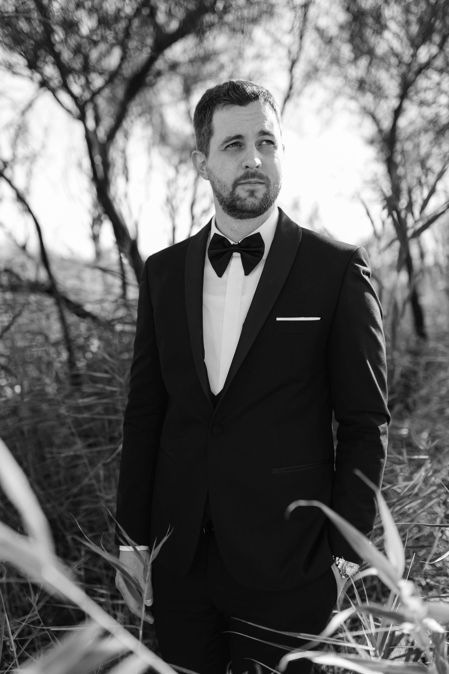 Black and white portrait of bearded groom in black tuxedo standing among trees outdoors.