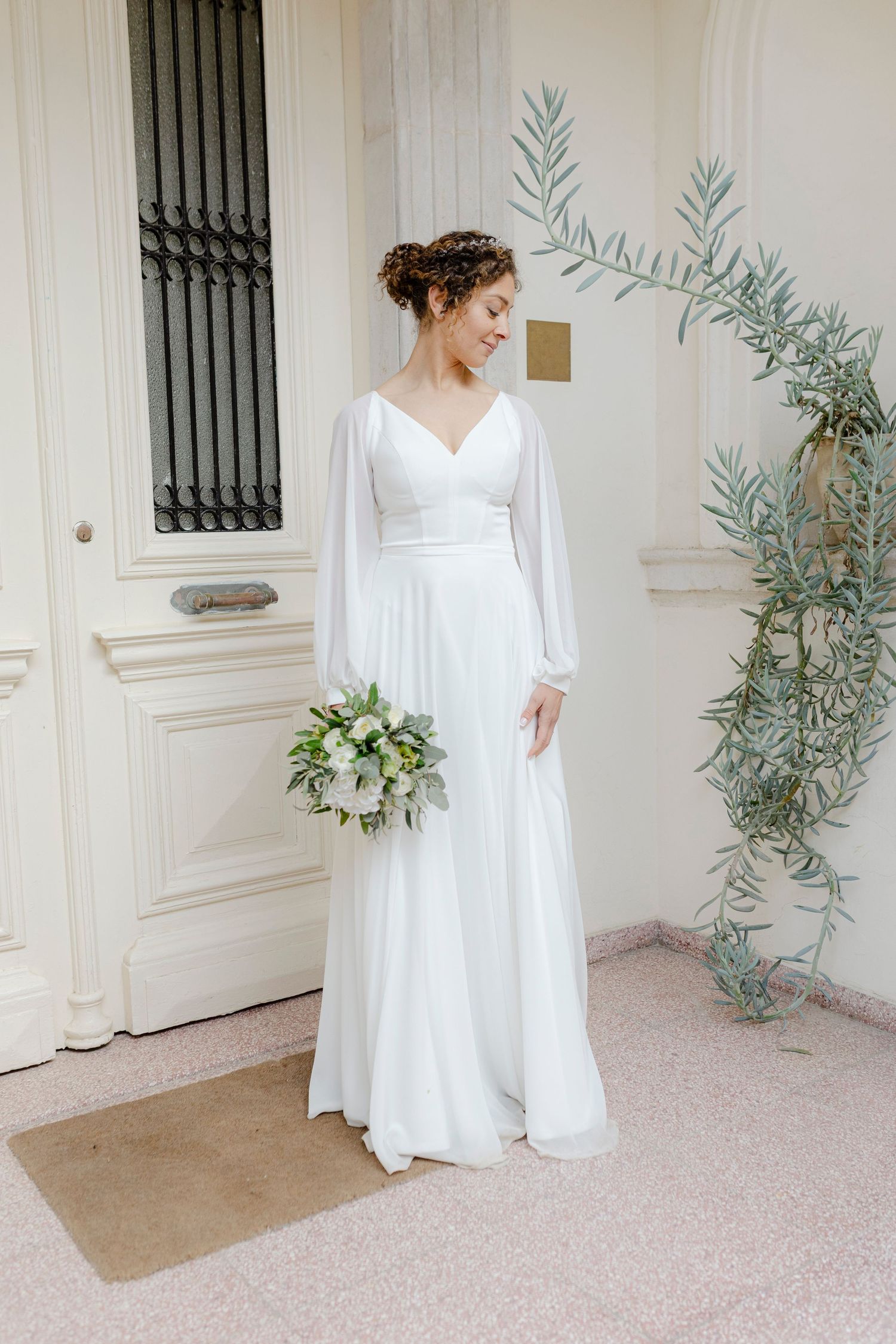 Bride in elegant white long-sleeve chiffon gown holding greenery bouquet, standing by ornate door with olive branch decor.