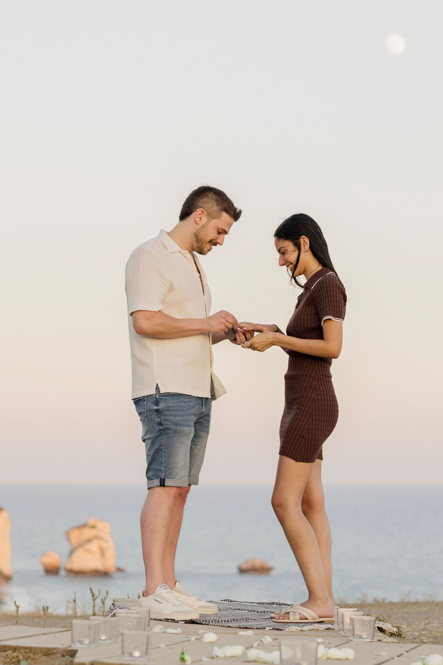 Man placing engagement ring on woman's finger during romantic beach proposal at sunset with ocean rocks in background.