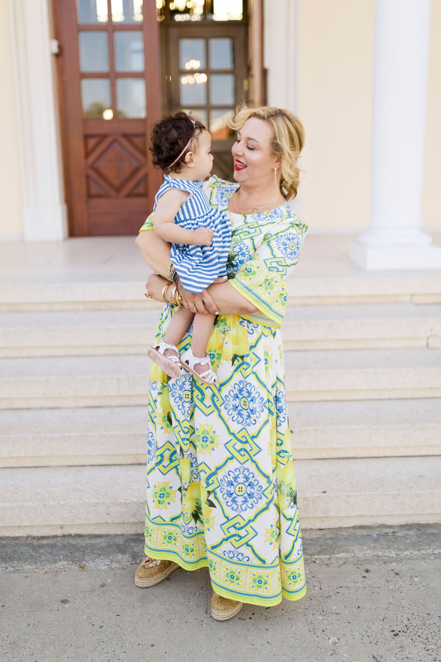 Woman in yellow floral maxi dress holding a baby in blue striped outfit on building steps.