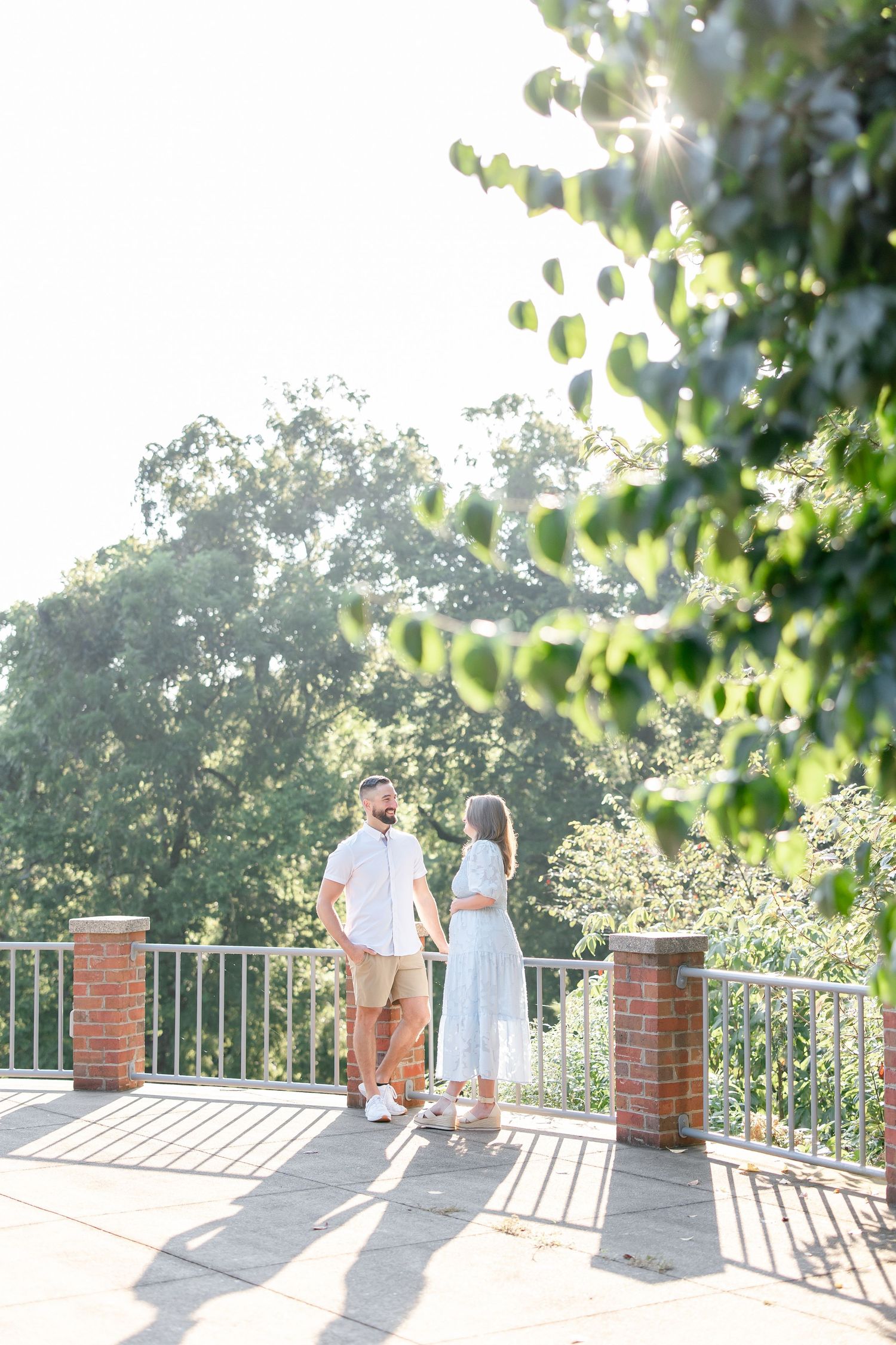 Couple stands together on a sunlit balcony with brick pillars surrounded by lush greenery at golden hour.