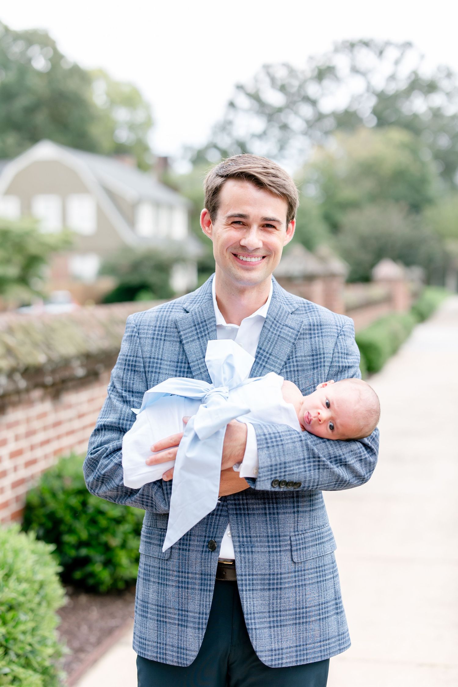 Man in checkered blazer holding swaddled baby on brick path outdoors.