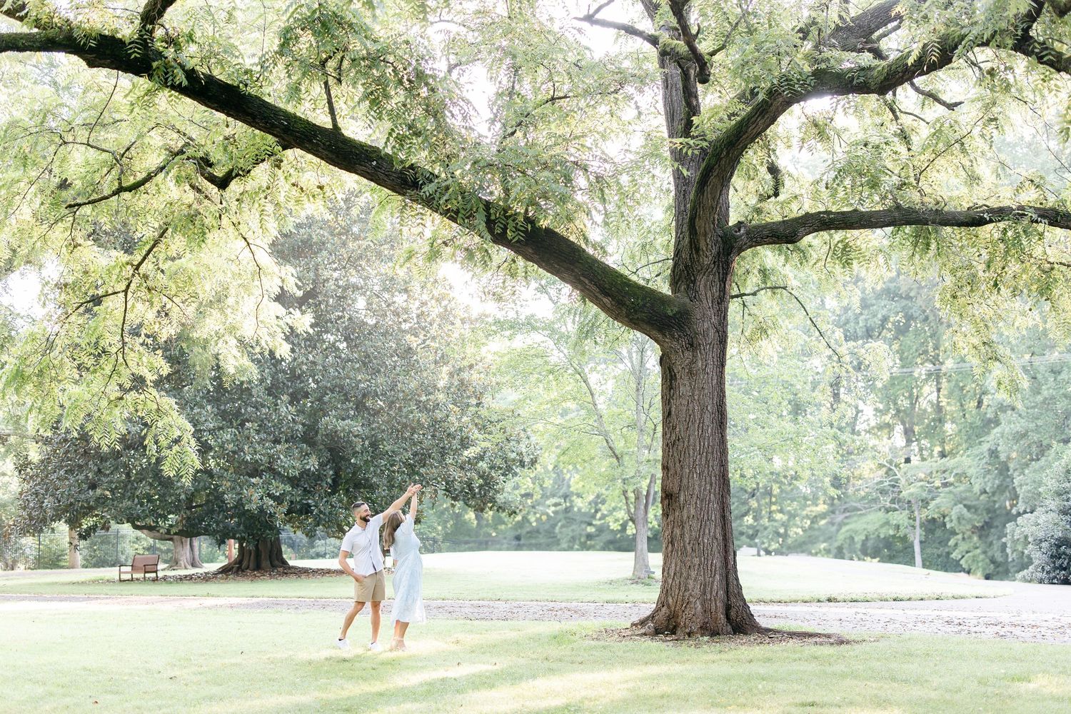 A couple swings from a low tree branch in a serene park setting during golden hour lighting.
