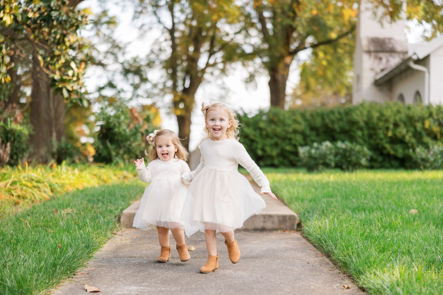 Two toddlers in matching white dresses walk hand in hand through a garden path during autumn.