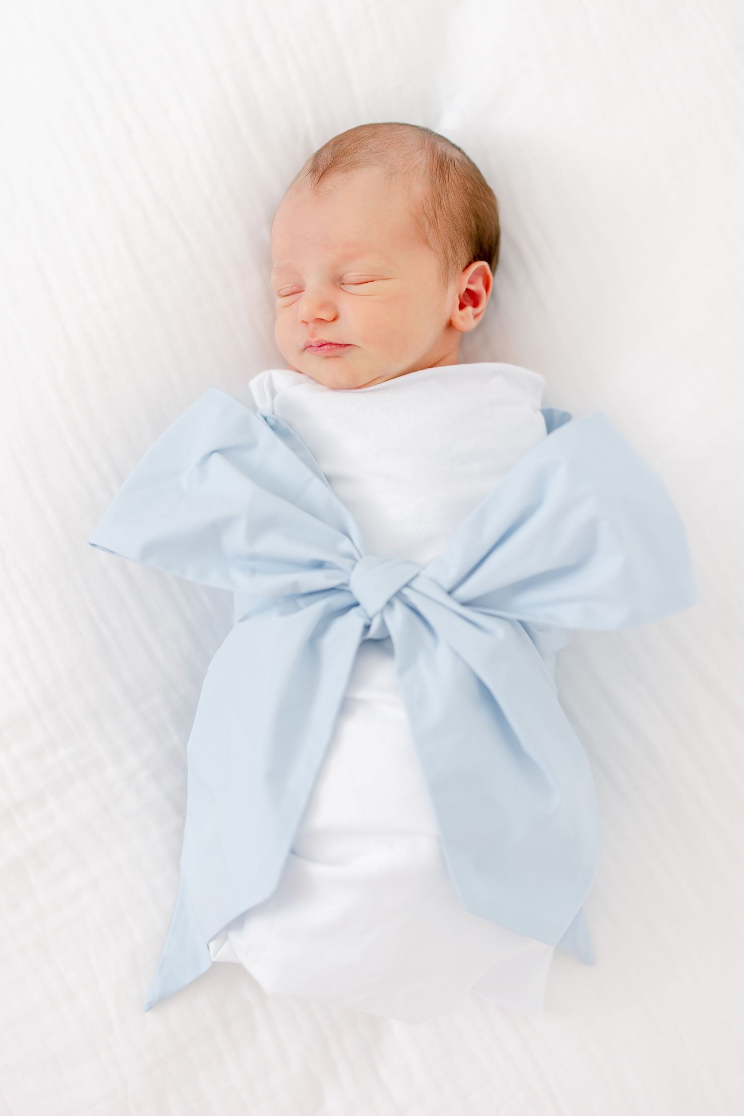 A sleeping baby swaddled in white with a large light blue bow wrapped around their middle on a white background.
