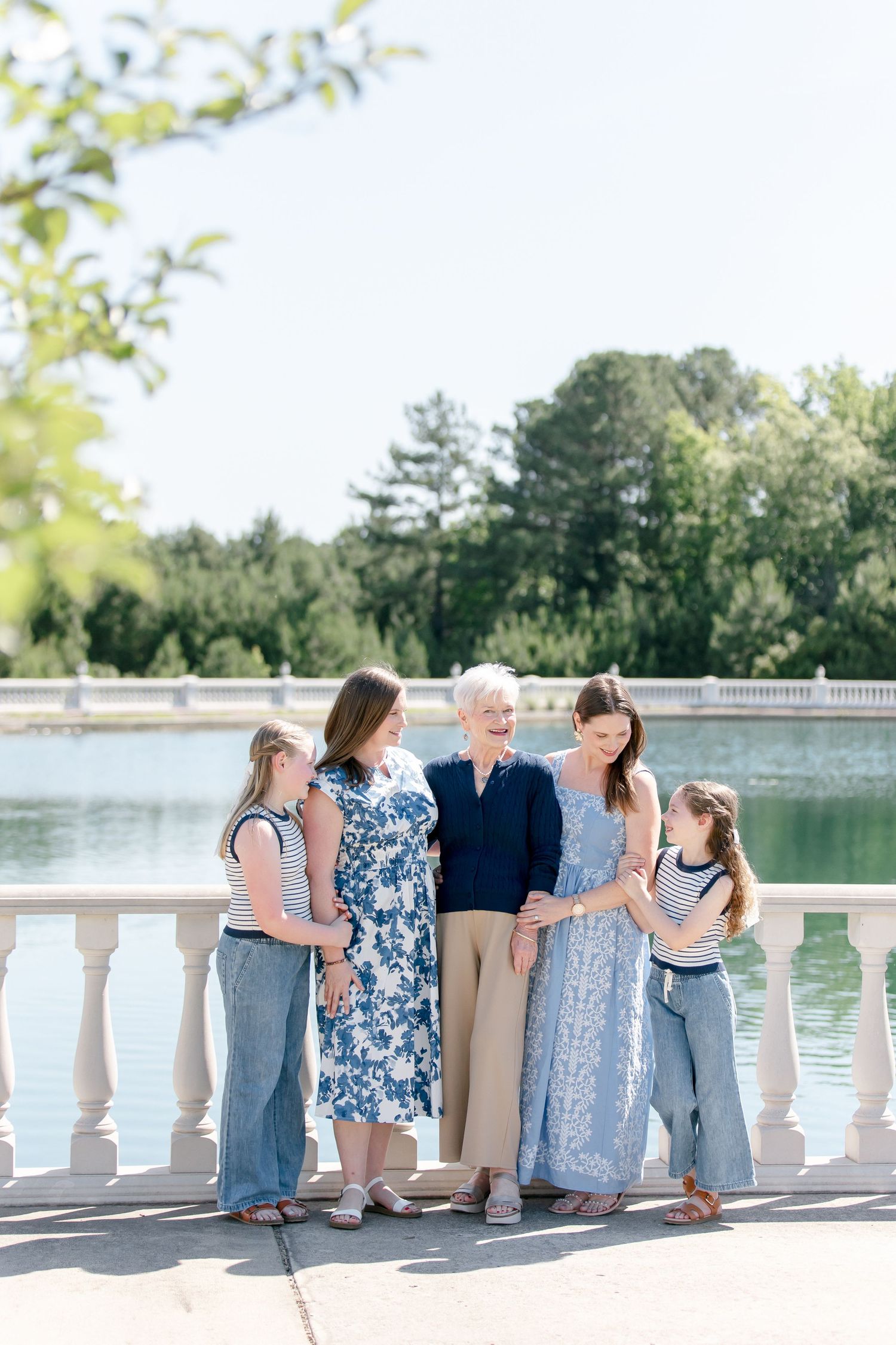 Family wearing coordinated blue and white outfits poses along waterfront stone railing on a sunny day.