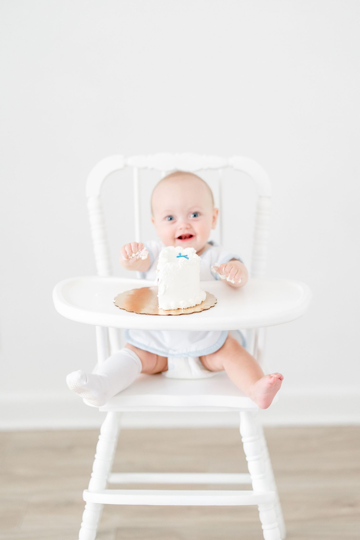 Smiling baby in high chair reaches for small white cake on wooden floor.