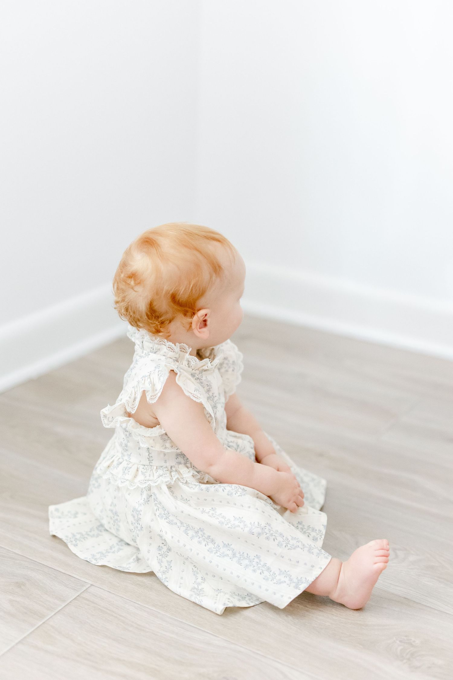 Baby wearing white lace dress lies on light wood floor in minimalist photography series.