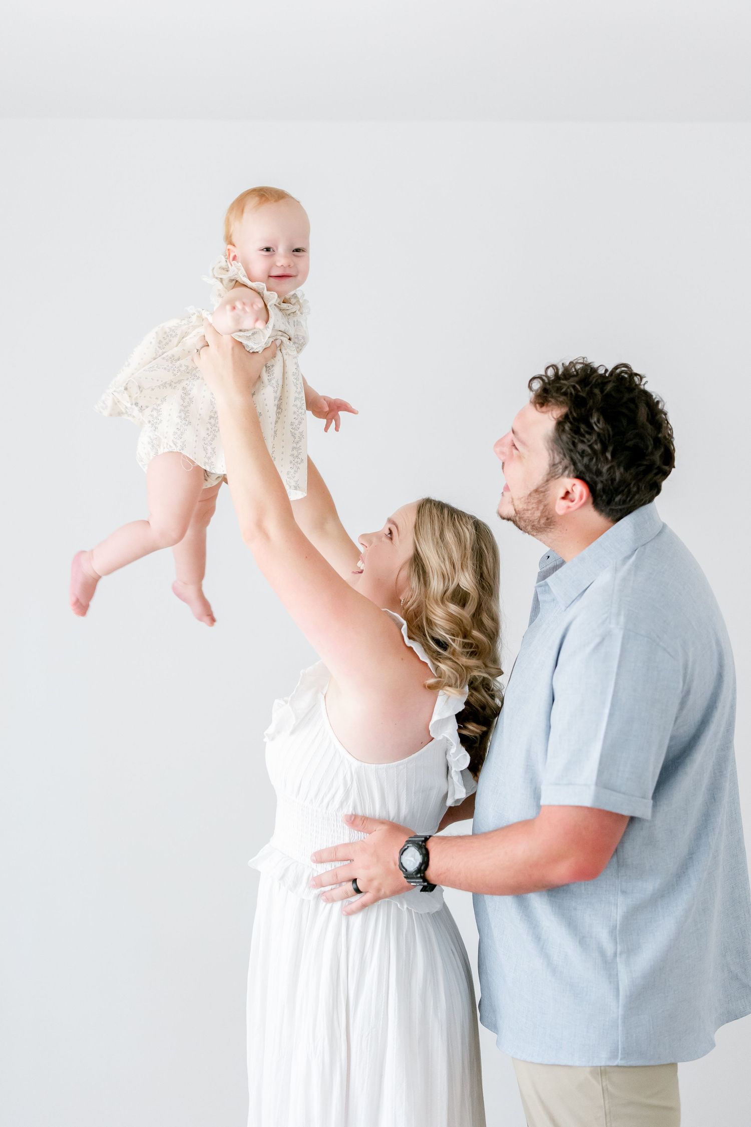 Parents in white and blue outfits lift their baby wearing a lace dress against a light gray studio backdrop.